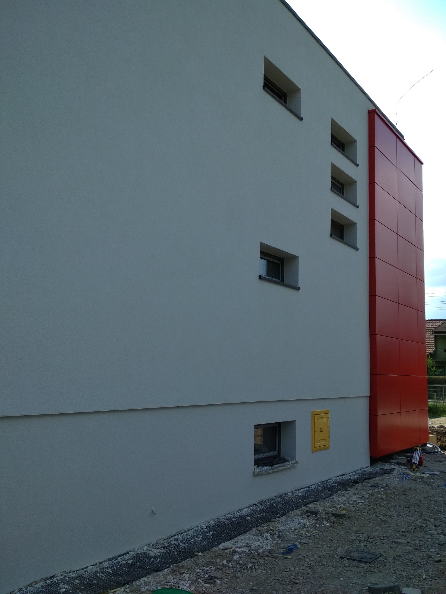 Nowoczesny dom z jasnoszarą elewacją i pionowym panelem z czerwonych płyt, widoczna skrzynka gazowa w kolorze żółtym.