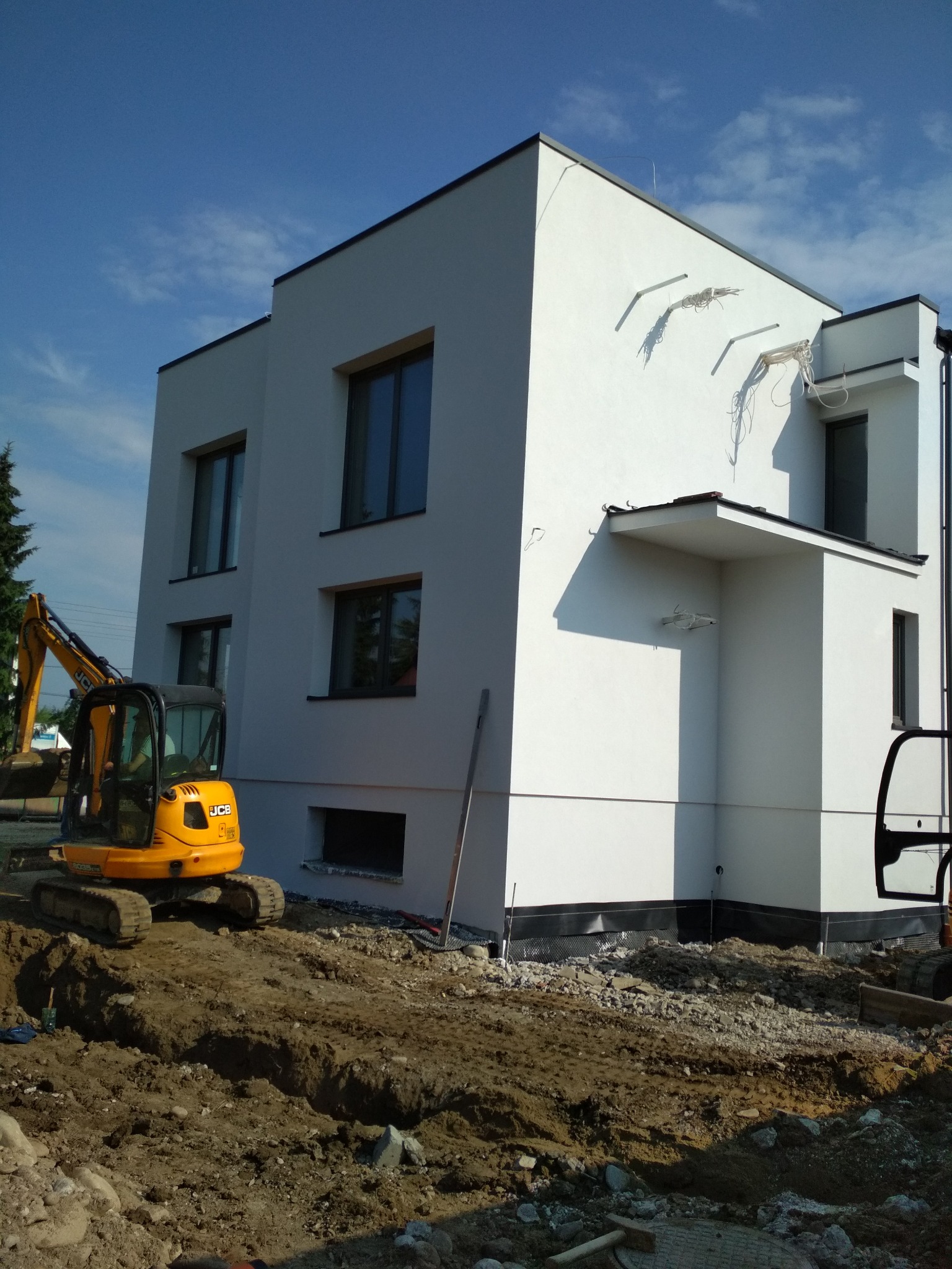 Nowoczesny, dwupiętrowy dom w trakcie budowy, widoczny żółty minikoparka JCB i przygotowany wykop pod fundamenty, na elewacji widoczne uchwyty do montażu ocieplenia.