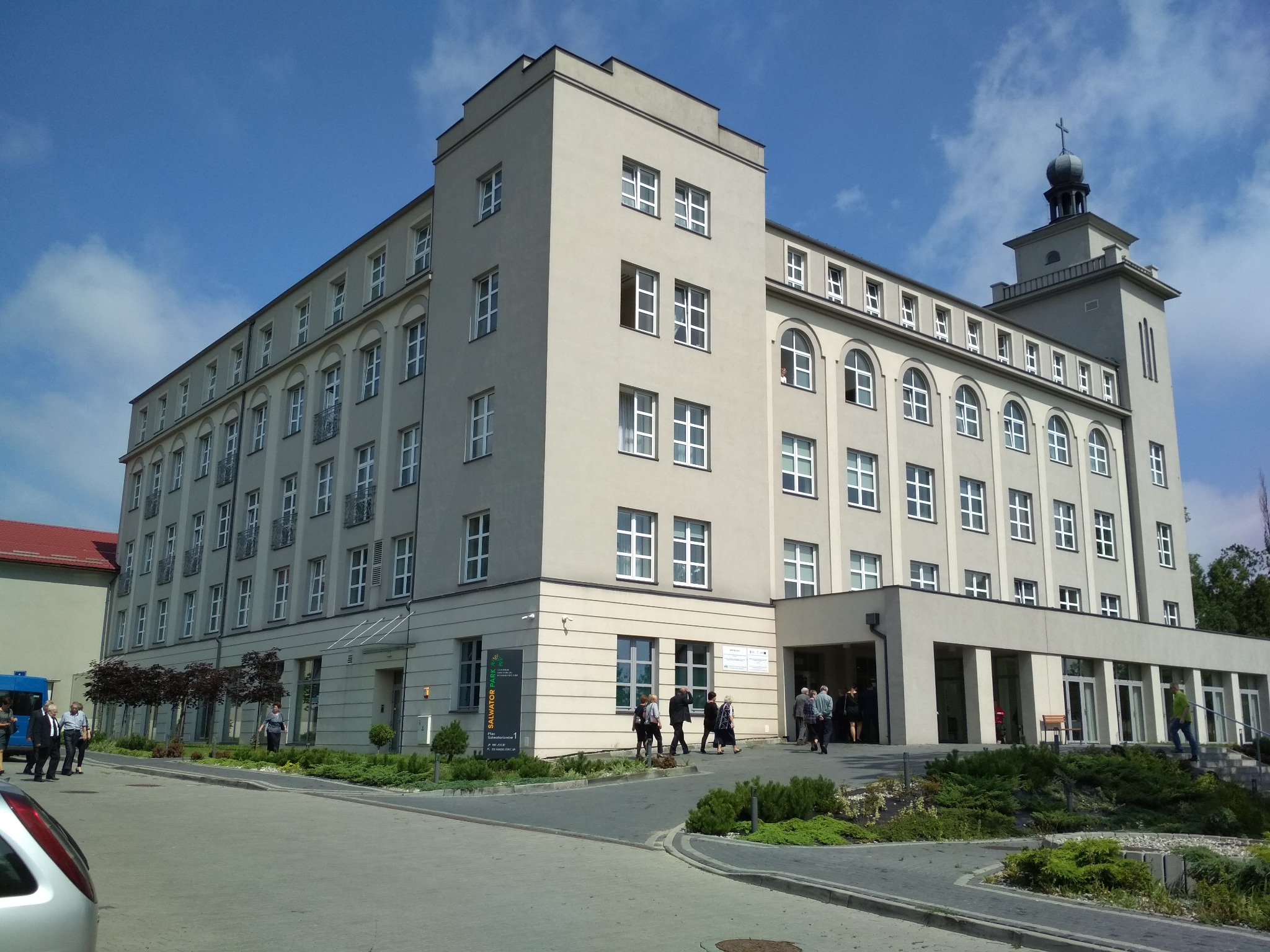 Duży, beżowy budynek z wieżą zwieńczoną krzyżem, widoczny z poziomu ulicy, z grupą ludzi wchodzących do głównego wejścia.