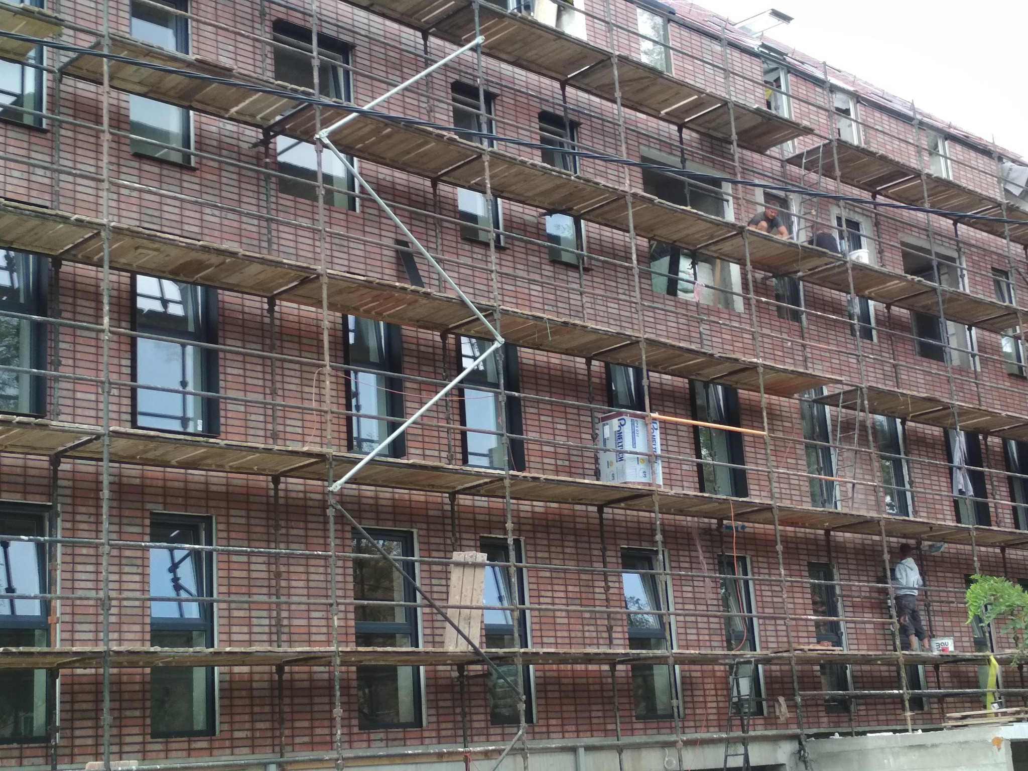 Murowany budynek wielorodzinny w trakcie prac elewacyjnych, widoczne rusztowania i pracownicy przy oknach.