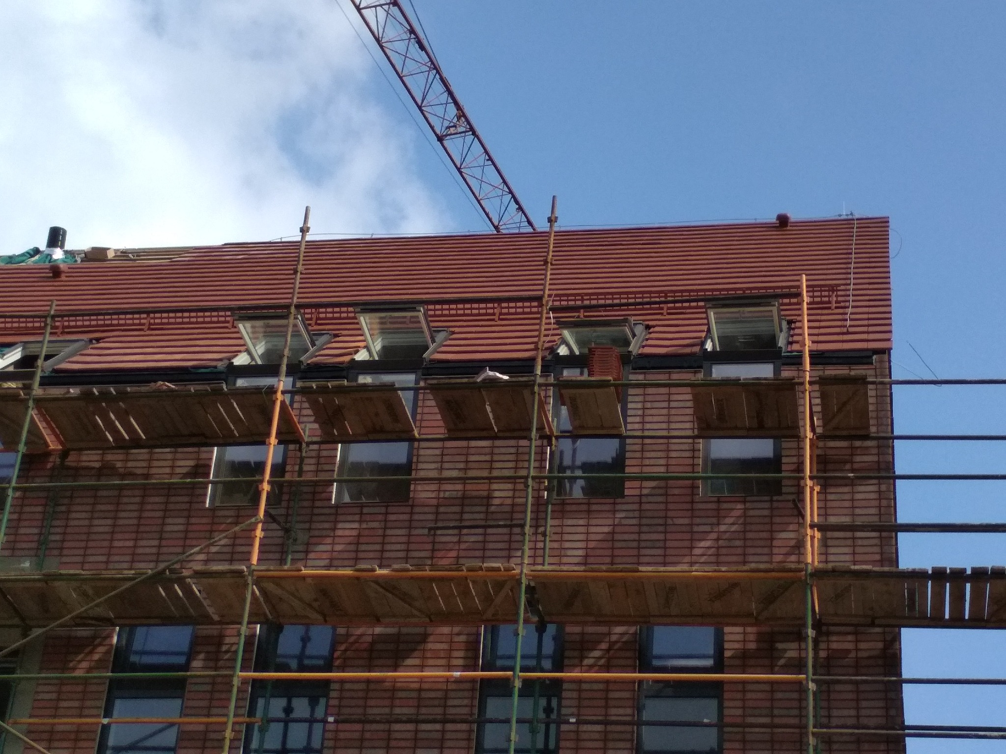 Wielopiętrowy budynek z czerwonej cegły w trakcie budowy, widoczne rusztowania, otwarte okna dachowe i dźwig budowlany na tle błękitnego nieba.