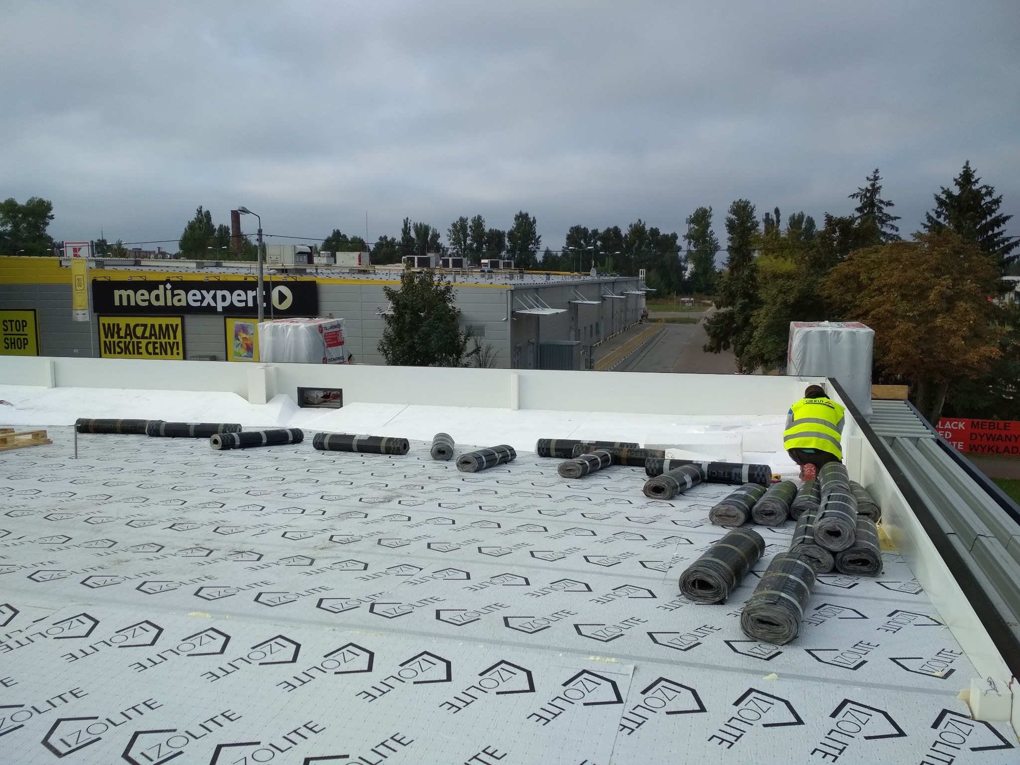 Prace na dachu budynku komercyjnego: rozłożona membrana izolacyjna z logo IzoLite, przygotowane rolki papy termozgrzewalnej i pracownik w kamizelce odblaskowej w trakcie pracy.