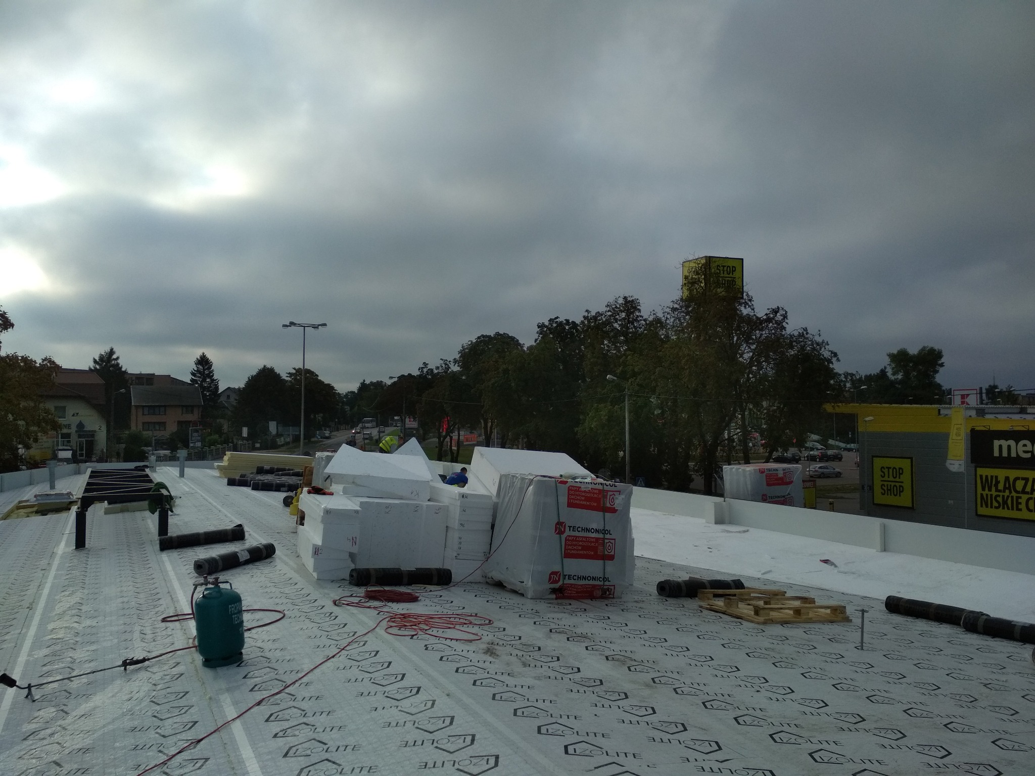 Prace izolacyjne na dachu płaskim: rozłożona membrana IZO-LITE z widocznymi zwojami papy, butla z gazem i przygotowane płyty izolacyjne TECHNONICOL, w tle budynki miejskie i pochmurne niebo.