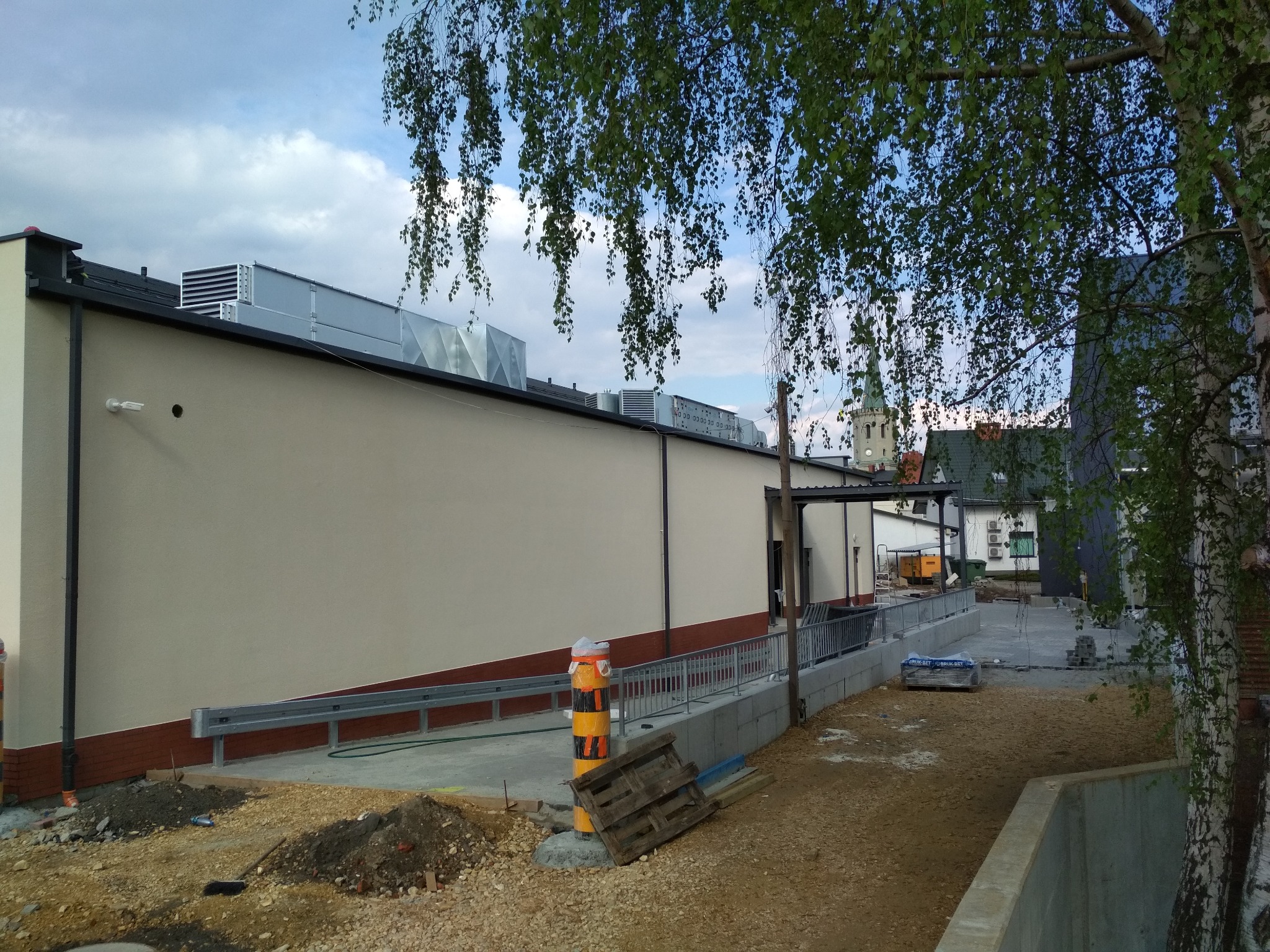 Narożne ujęcie budynku z ocieploną elewacją w kolorze beżowym, widoczne elementy wentylacji na dachu, barierki ochronne, teren w trakcie zagospodarowania, w tle fragment wieży kościelnej.