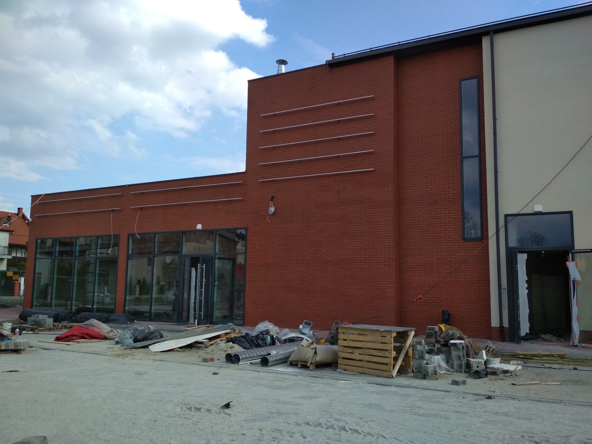 Budynek w trakcie prac wykończeniowych: czerwona cegła, duże okna, widoczne materiały budowlane i narzędzia.