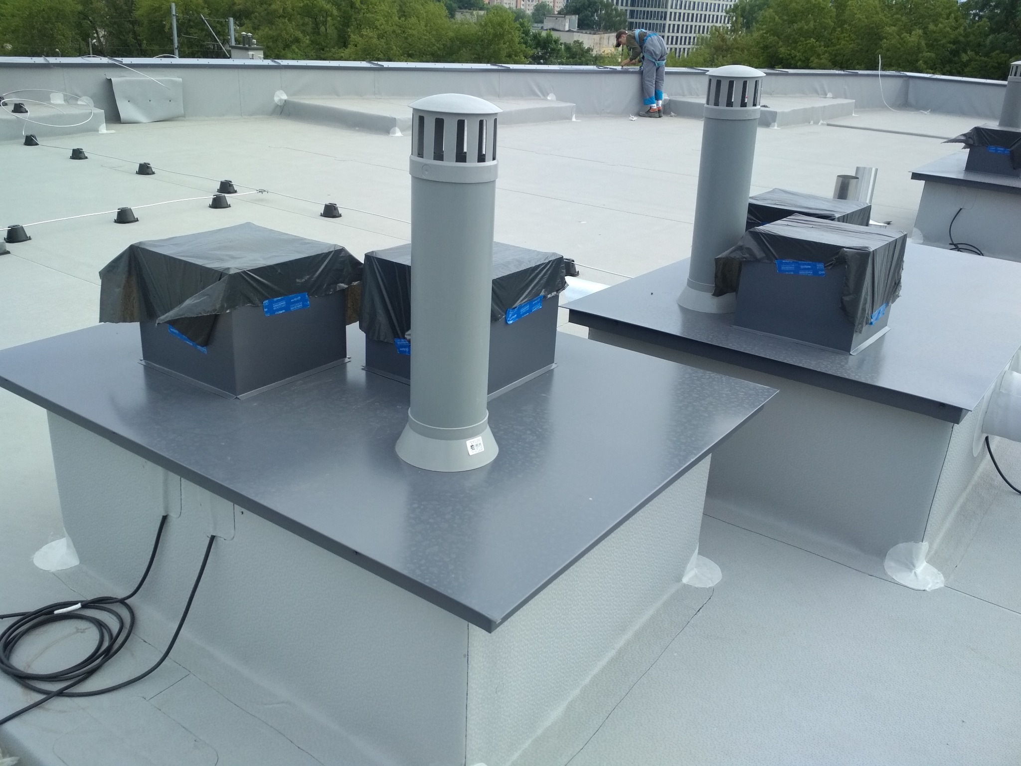 Płaski dach pokryty szarą membraną z kilkoma szarymi kominami wentylacyjnymi i osłonami na urządzeniach, pracownik w oddali wykonuje prace montażowe.