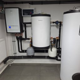CZUPALLA - Instalacja dwóch zbiorników akumulacyjnych Vaillant, pompy ciepła i grzejnika w kotłowni z szarymi płytkami podłogowymi i czarnymi rurami.