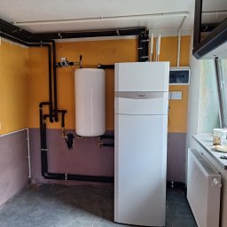 CZUPALLA - Biała pompa ciepła Vaillant z podłączonym zasobnikiem ciepłej wody, rury instalacji biegnące wzdłuż ściany w pomieszczeniu z pomalowanymi ścianami i szarą podłogą z płytek.