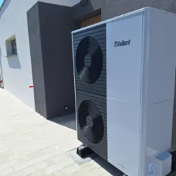 Biała pompa ciepła Vaillant aroTHERM zamontowana na szarym bruku przed nowoczesnym budynkiem z dwukolorową elewacją (biało-szarą).