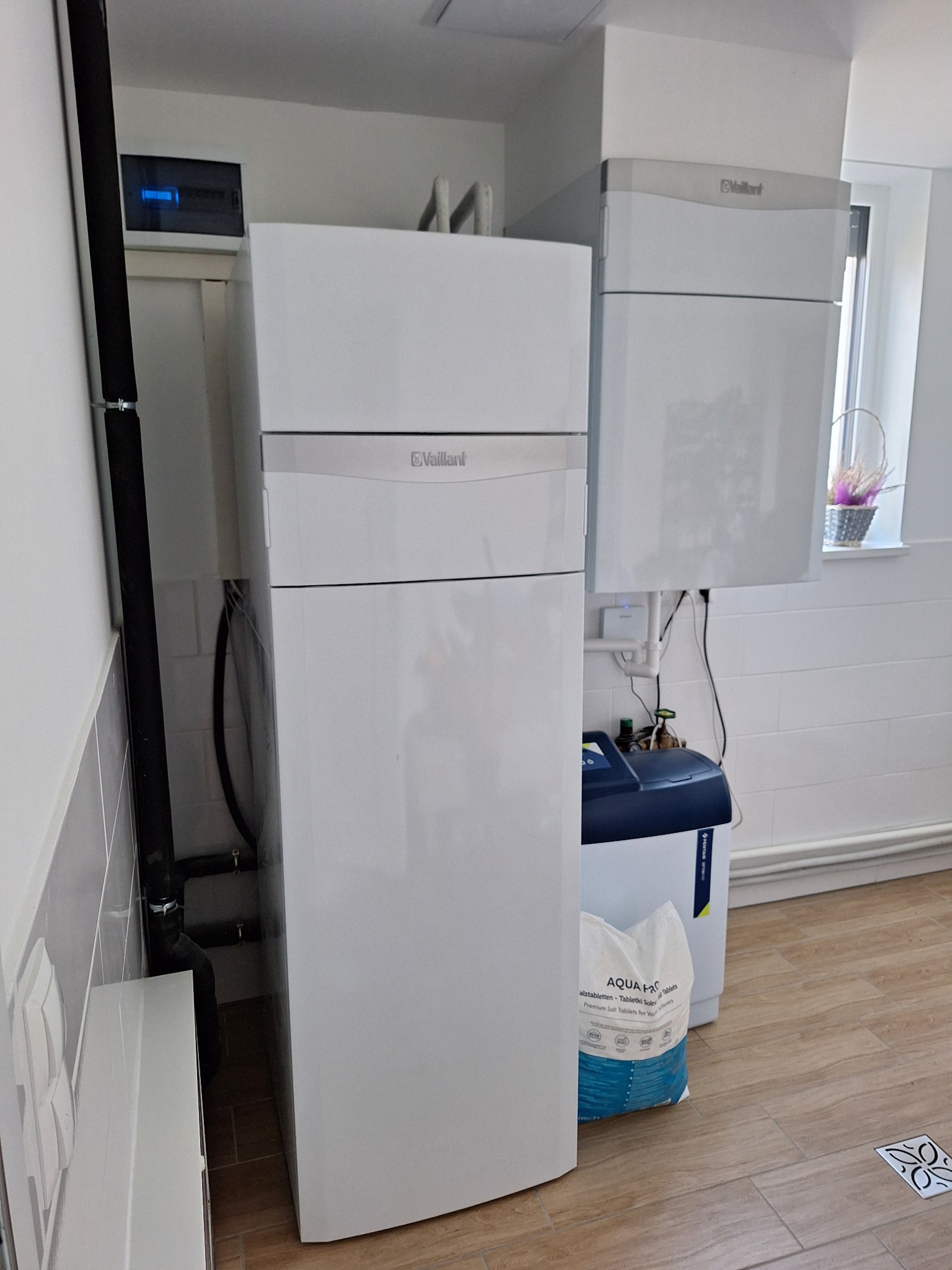 Biała instalacja grzewcza marki Vaillant z podłączonymi rurami, obok zmiękczacz wody i worek soli tabletkowanej na podłodze z płytek imitujących drewno.