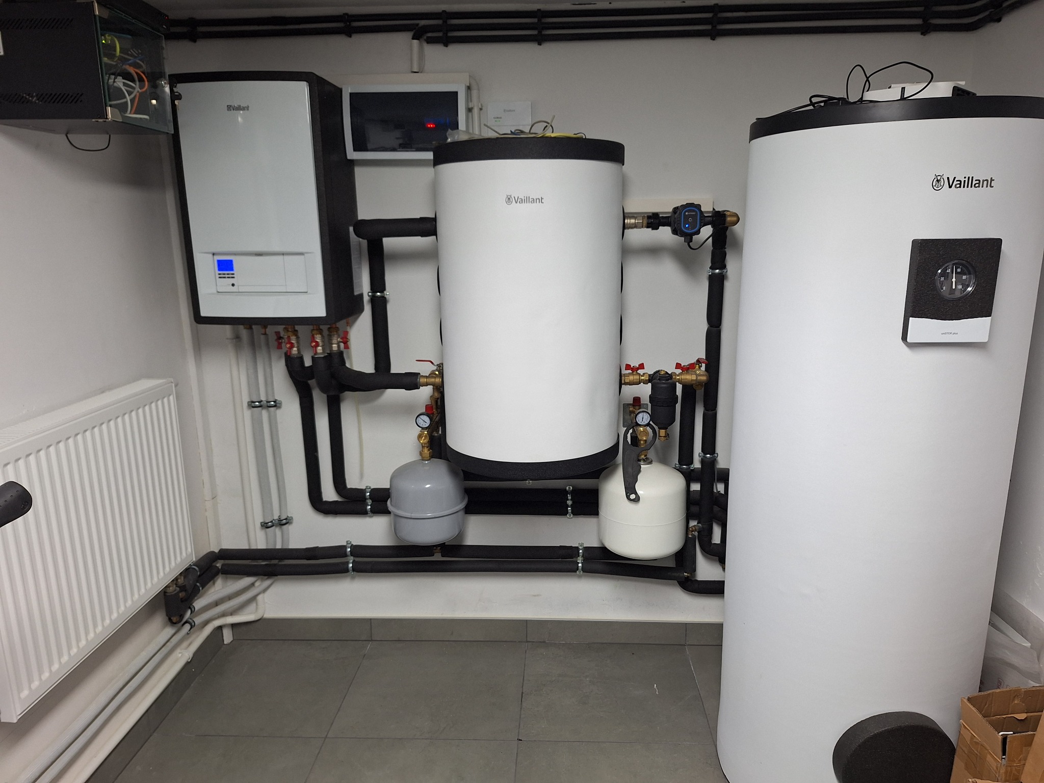 Instalacja dwóch zbiorników akumulacyjnych Vaillant, pompy ciepła i grzejnika w kotłowni z szarymi płytkami podłogowymi i czarnymi rurami.