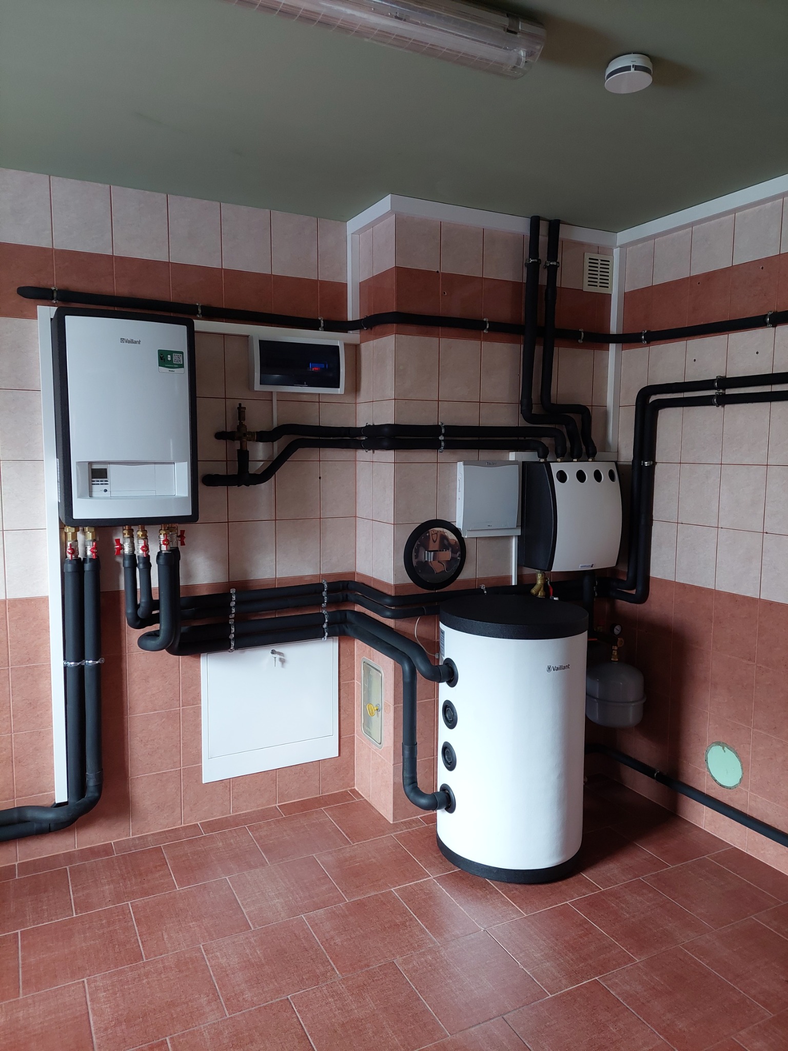 Zestaw urządzeń grzewczych marki Vaillant w pomieszczeniu wyłożonym kafelkami w odcieniach brązu, widoczne: wiszący piec, zasobnik ciepłej wody i inne elementy instalacji.