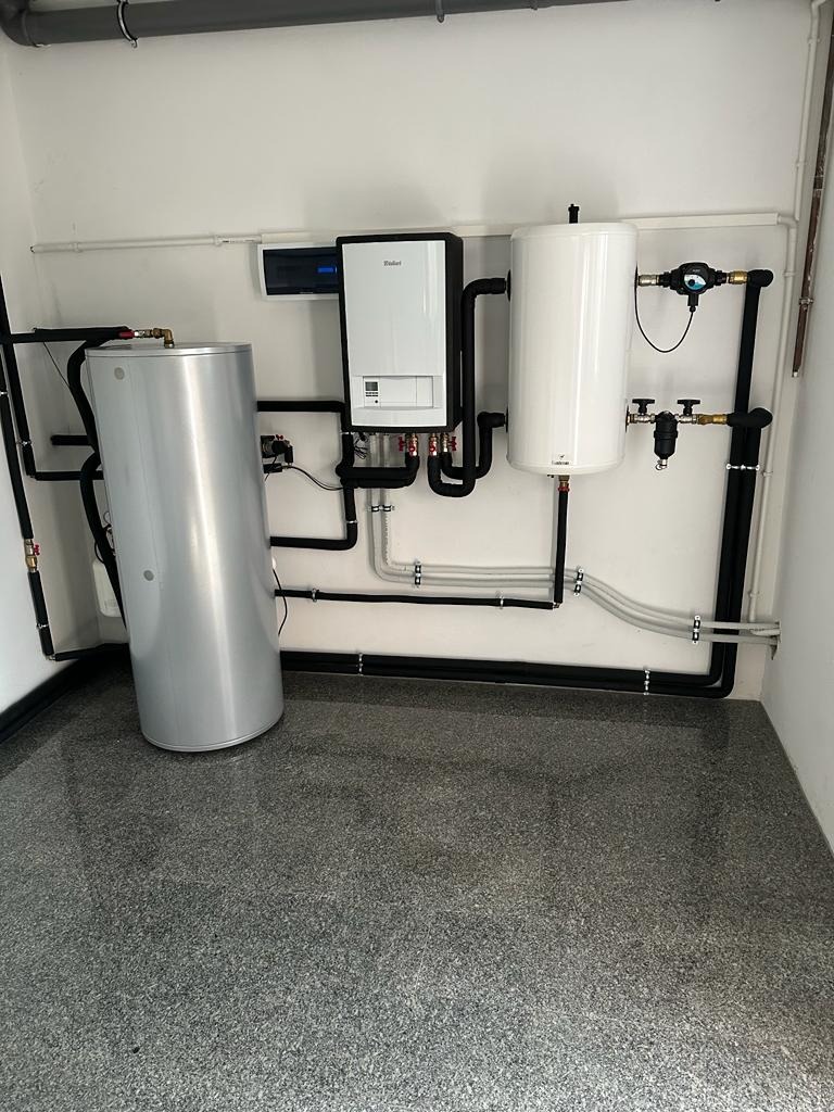 Zestaw urządzeń grzewczych Vaillant: srebrny zasobnik CWU, biały kocioł grzewczy i biały zbiornik buforowy, połączone czarnymi rurami w kotłowni z szarą posadzką z żywicy.