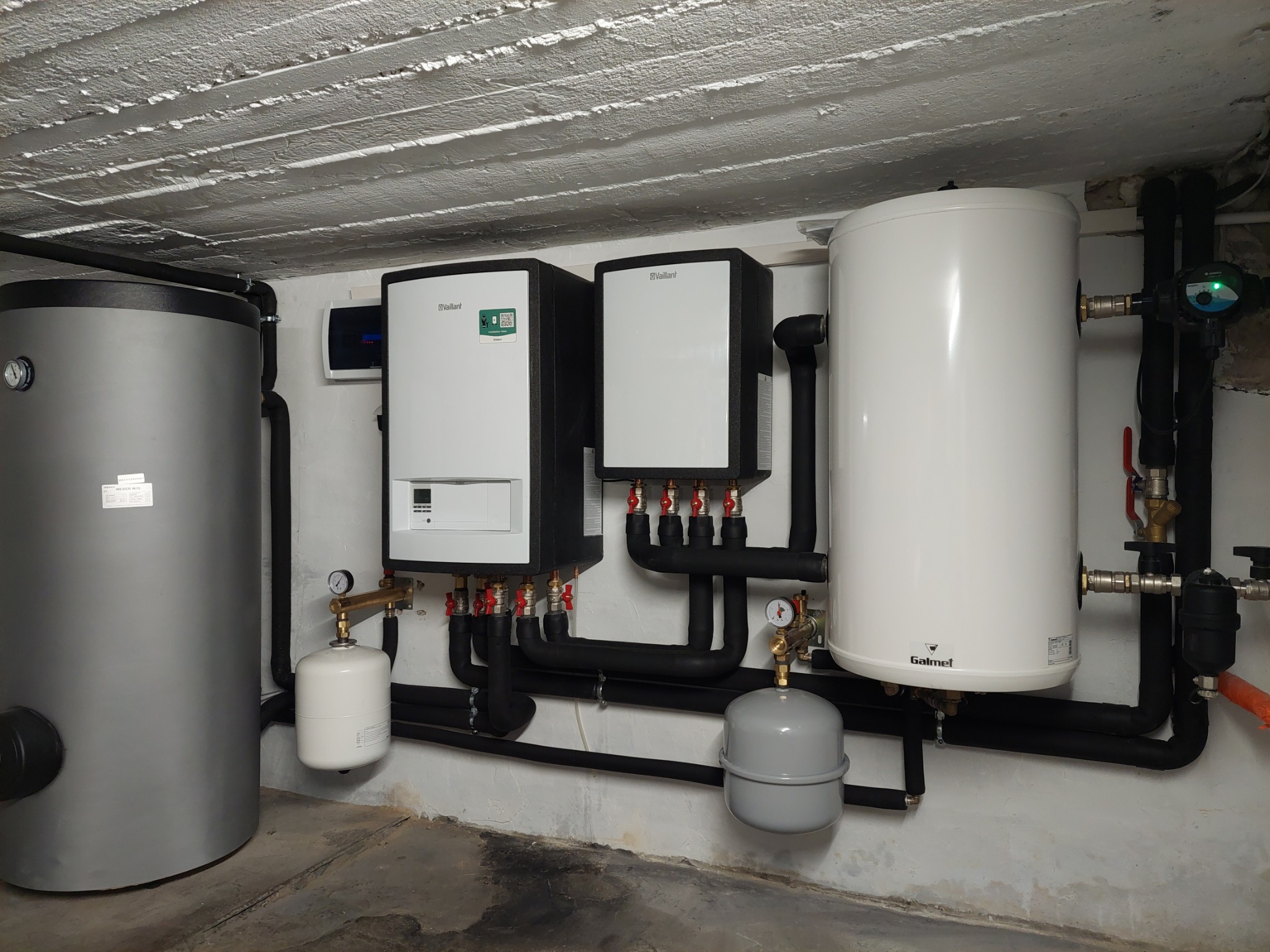 Zestaw urządzeń grzewczych Vaillant i Galmet w piwnicy: dwa piece, dwa zbiorniki, rury w izolacji, manometry, zawory. Widoczna surowa faktura betonowego stropu.