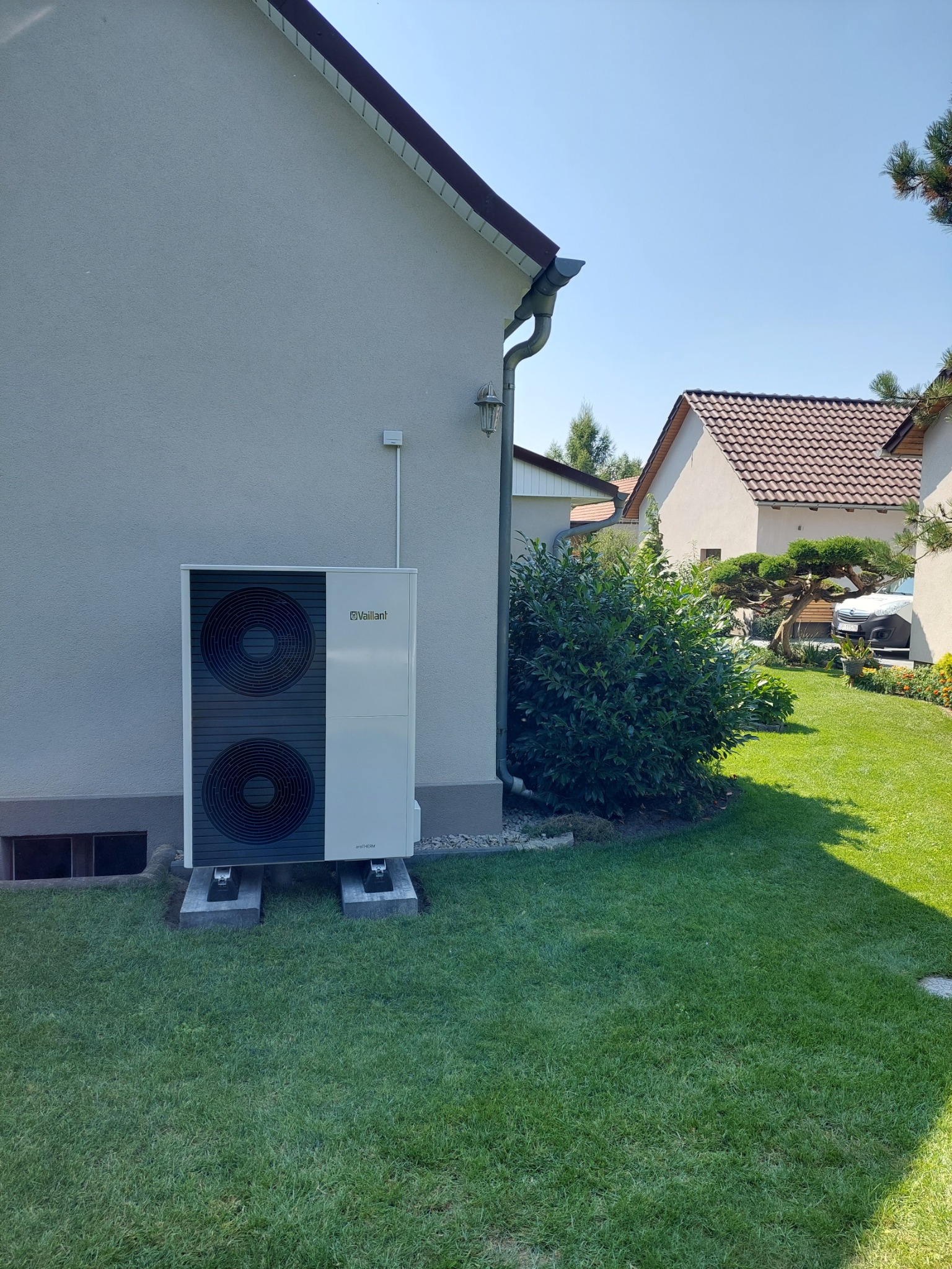 Zewnętrzna pompa ciepła Vaillant aroTHERM zamontowana na betonowych podstawach przy ścianie domu z trawnikiem w słoneczny dzień.