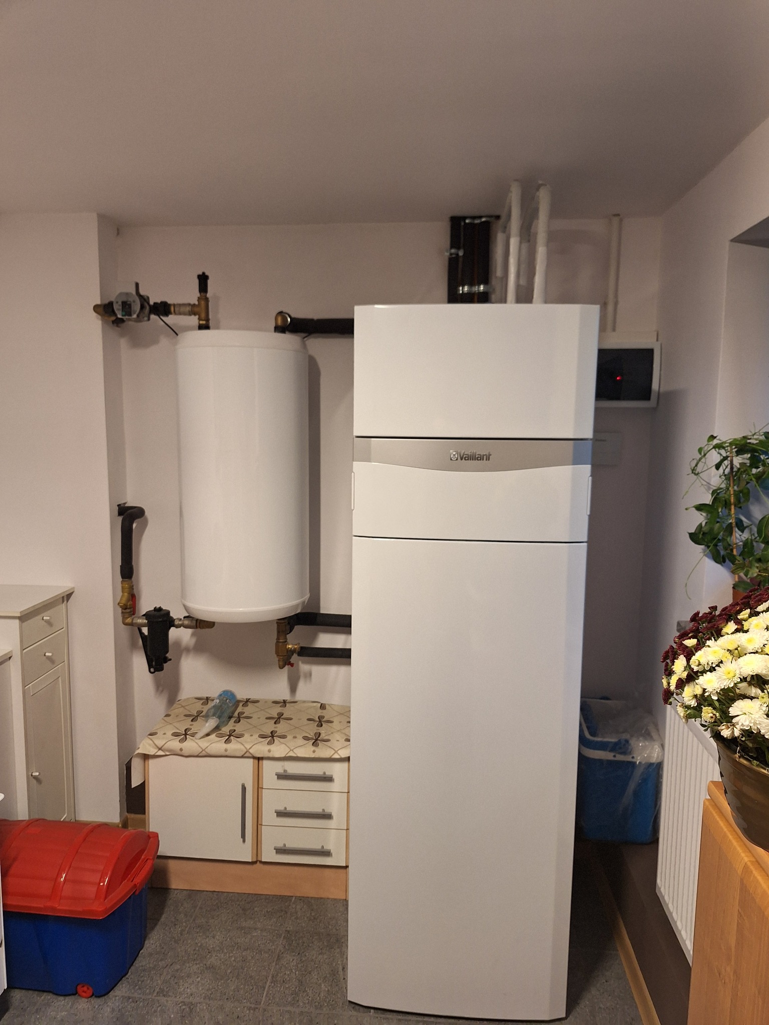 Biała pompa ciepła Vaillant z zasobnikiem ciepłej wody w kotłowni, widoczne orurowanie, armatura i sterownik na ścianie. Obok stoi szafka z przykryciem i skrzynka na narzędzia.