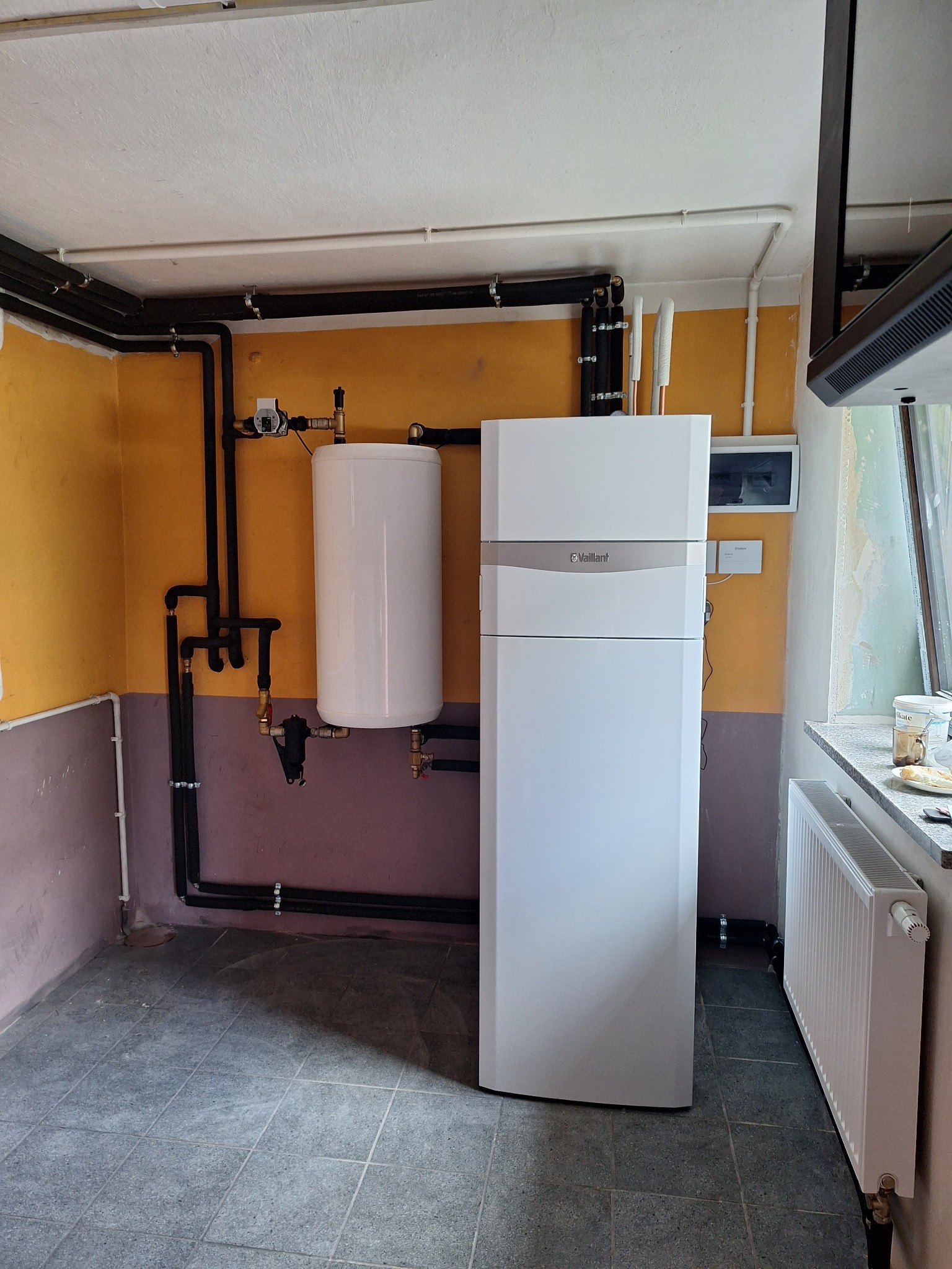 Biała pompa ciepła Vaillant z podłączonym zasobnikiem ciepłej wody, rury instalacji biegnące wzdłuż ściany w pomieszczeniu z pomalowanymi ścianami i szarą podłogą z płytek.