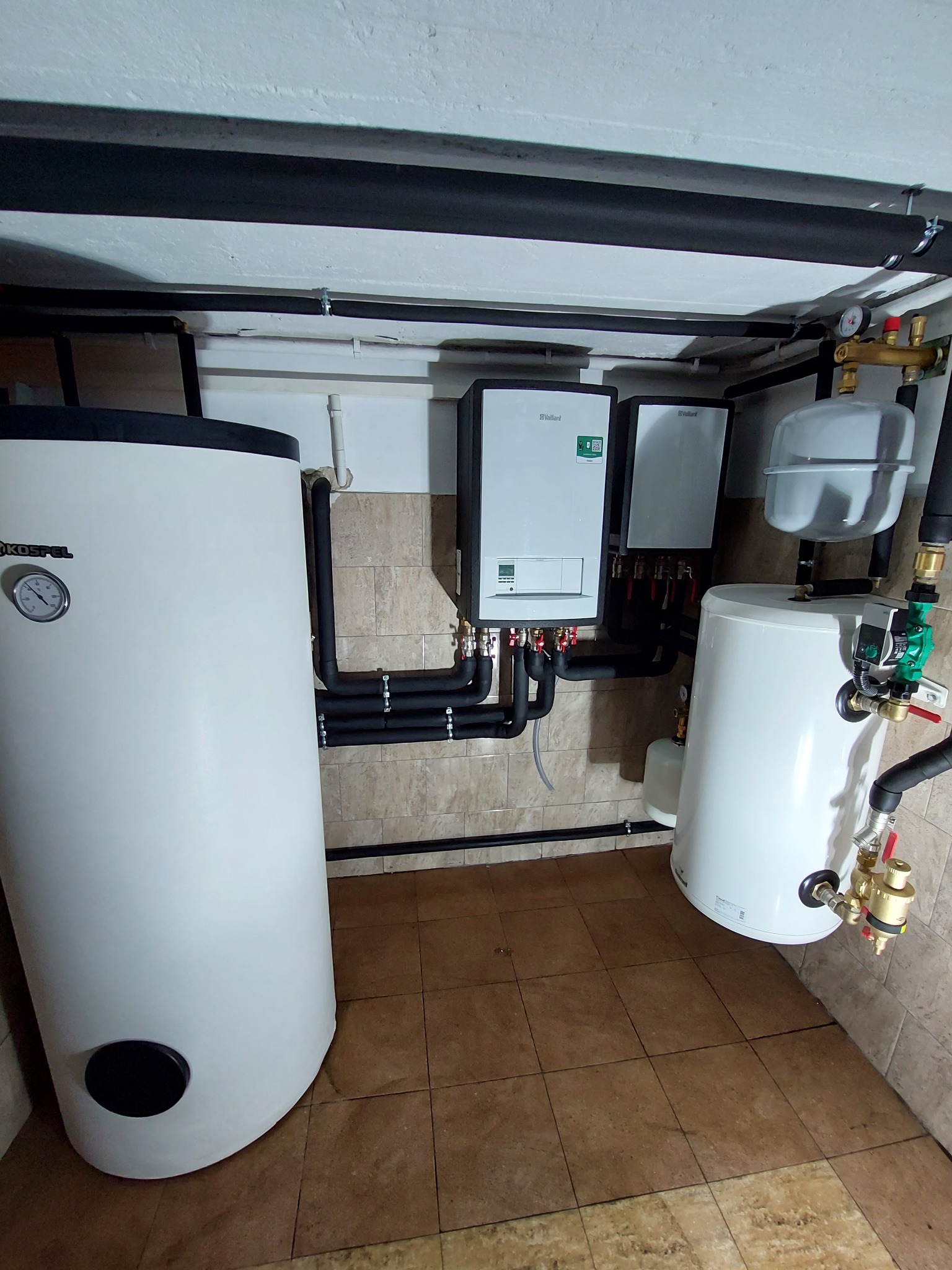 Kompletna instalacja hydrauliczna z dwoma zbiornikami (Kospel i Vaillant) oraz rurami w piwnicy wyłożonej brązowymi płytkami.