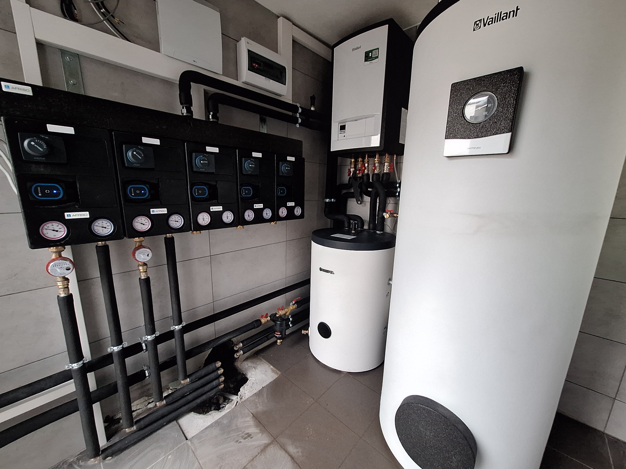 Instalacja kotłowni z dwoma zbiornikami, rozdzielaczami AFRISO i orurowaniem w pomieszczeniu z szarą glazurą.