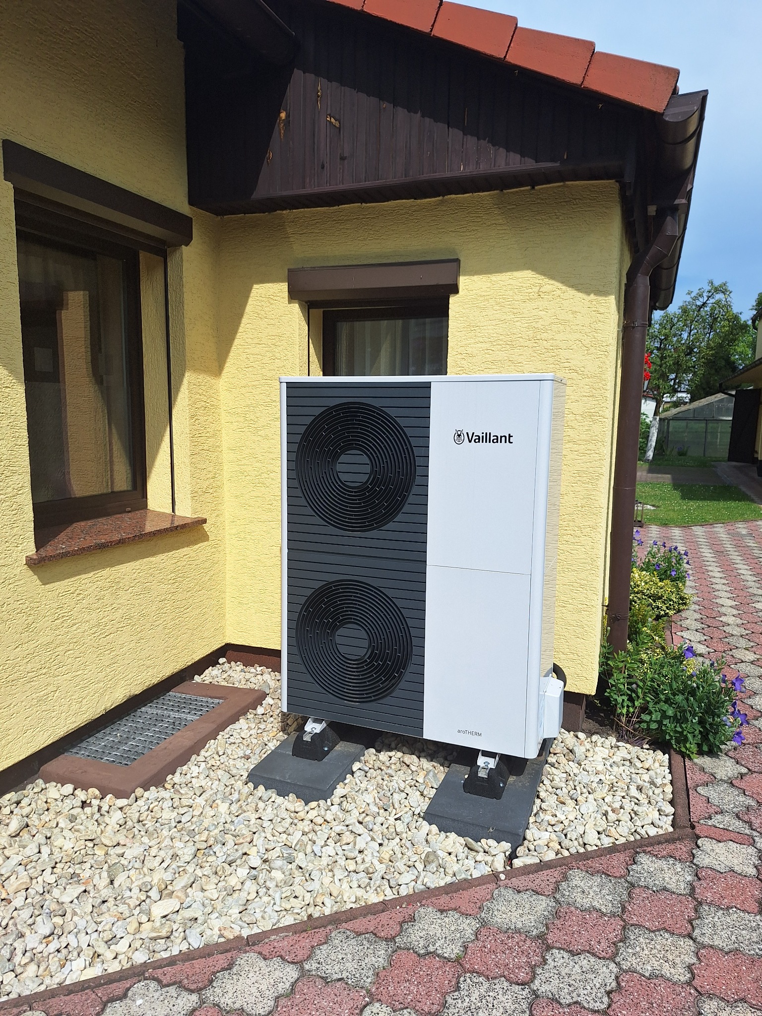Biało-szara jednostka zewnętrzna z dwoma wentylatorami marki Vaillant zainstalowana przy żółtej ścianie domu, na betonowych podstawach, otoczona białymi kamieniami i kostką brukową.