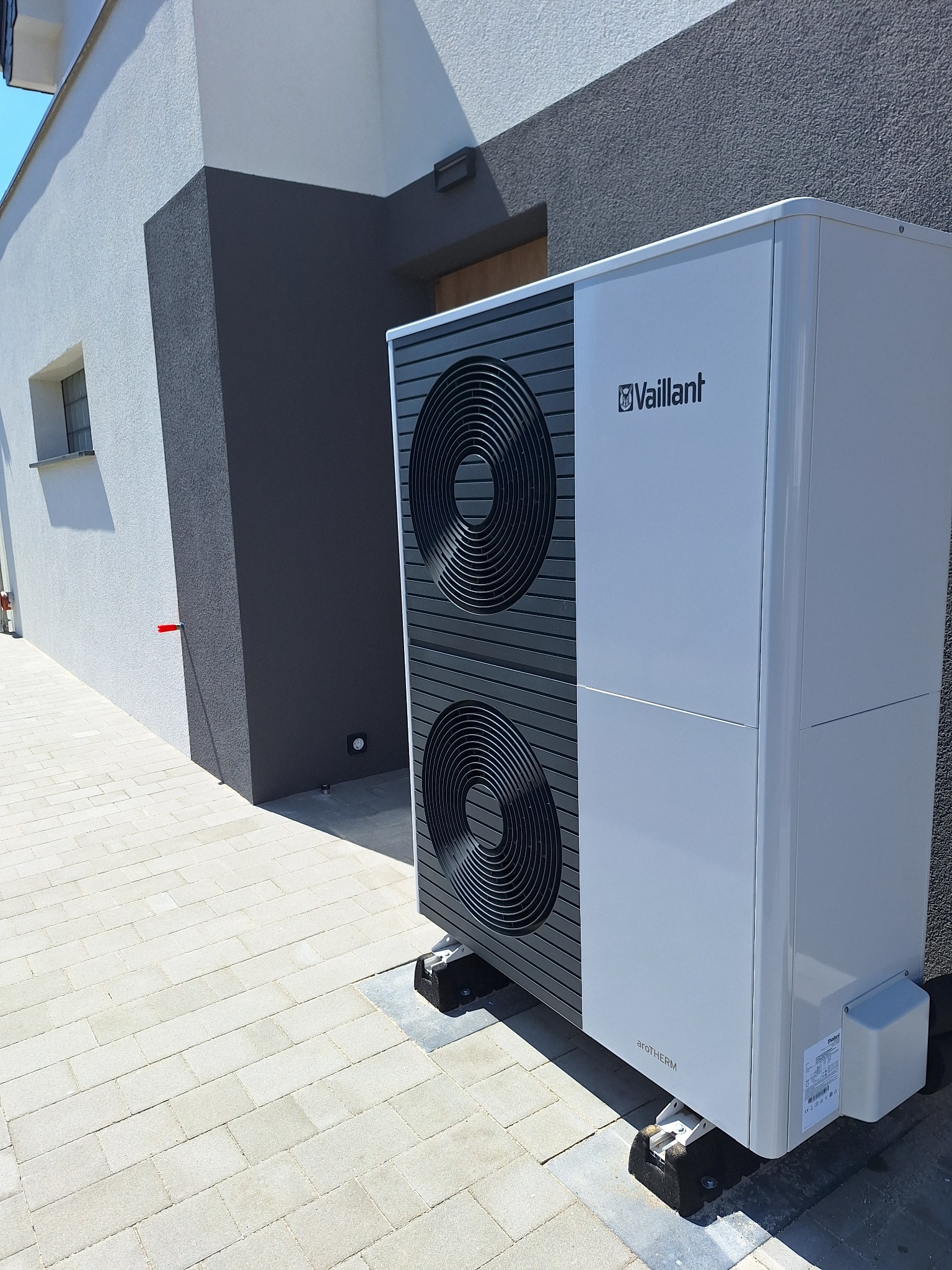 Biała pompa ciepła Vaillant aroTHERM zamontowana na szarym bruku przed nowoczesnym budynkiem z dwukolorową elewacją (biało-szarą).