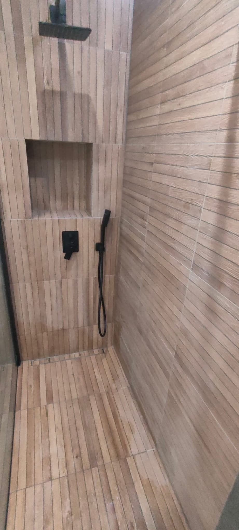 Nowoczesna kabina prysznicowa wykończona płytkami imitującymi drewno, wyposażona w deszczownicę i słuchawkę prysznicową w kolorze czarnym, z wnęką na kosmetyki w ścianie.