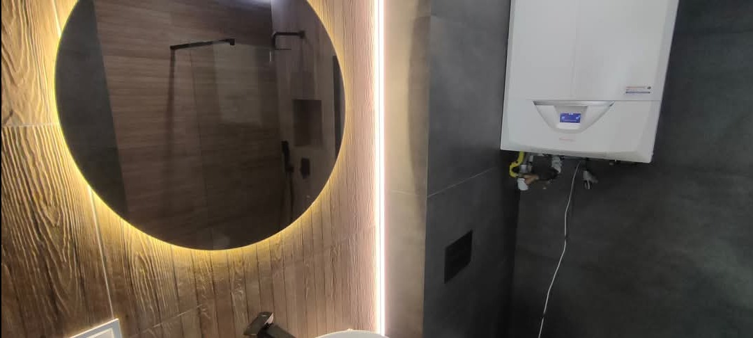 Nowoczesna łazienka z okrągłym lustrem podświetlanym LED, brązowymi płytkami imitującymi drewno i szarymi płytkami na ścianie, z widocznym piecem gazowym.