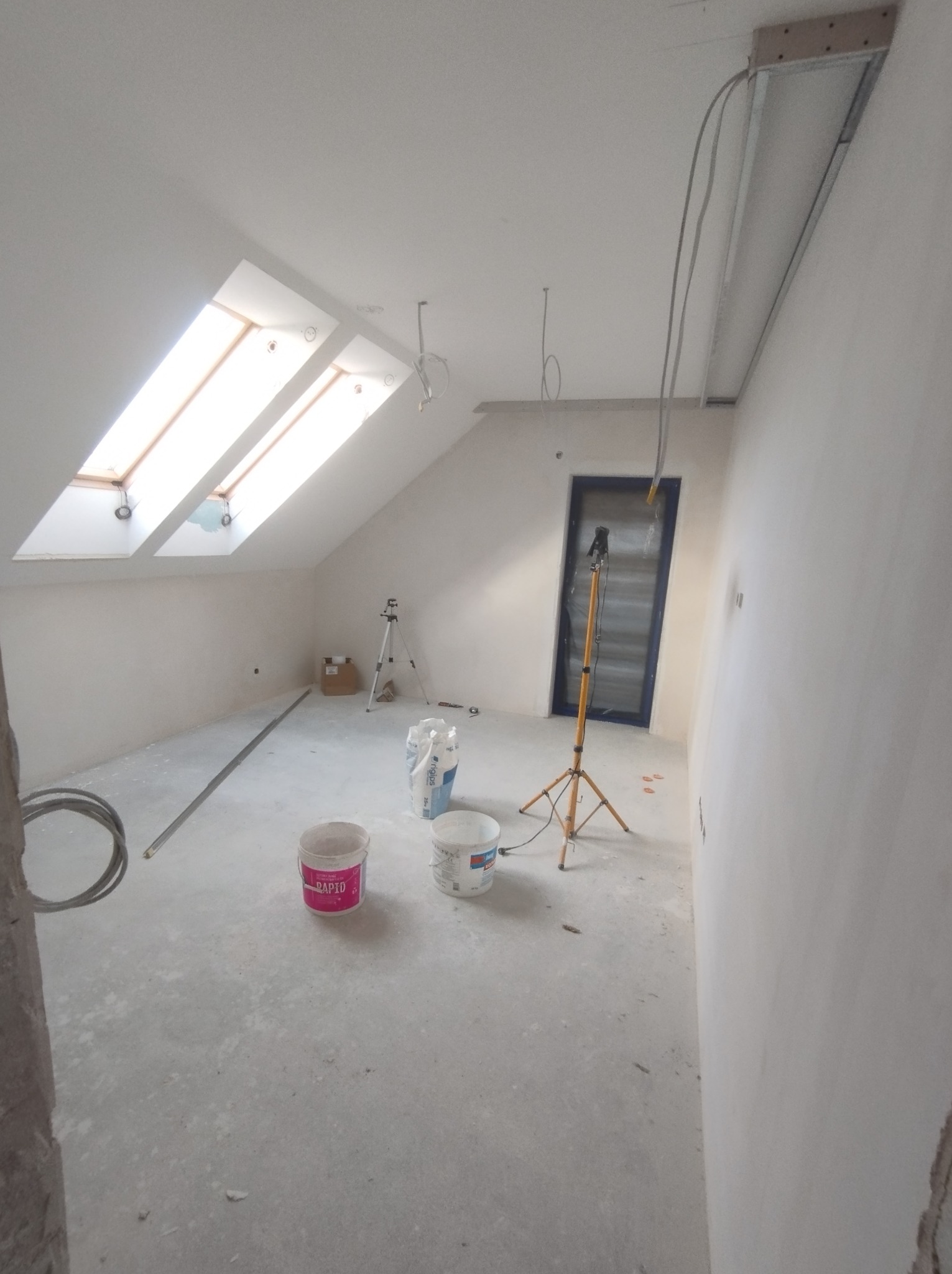 Pokój na poddaszu w trakcie remontu: widoczne okno dachowe, worki z gipsem, wiaderka z zaprawą, lampa na statywie, przygotowane przewody elektryczne.