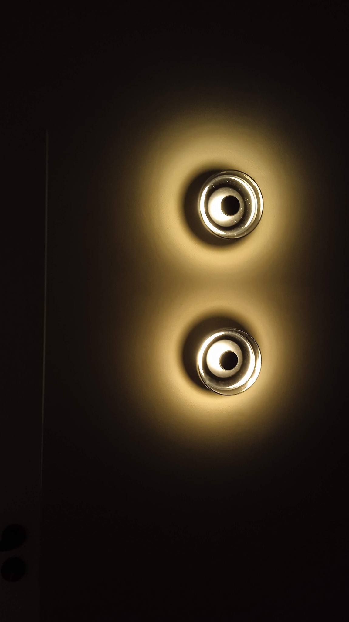 Dwa okrągłe, szklane kinkiety ścienne z ciepłym światłem, umieszczone pionowo na ciemnej ścianie, tworzące okrągłe plamy światła.