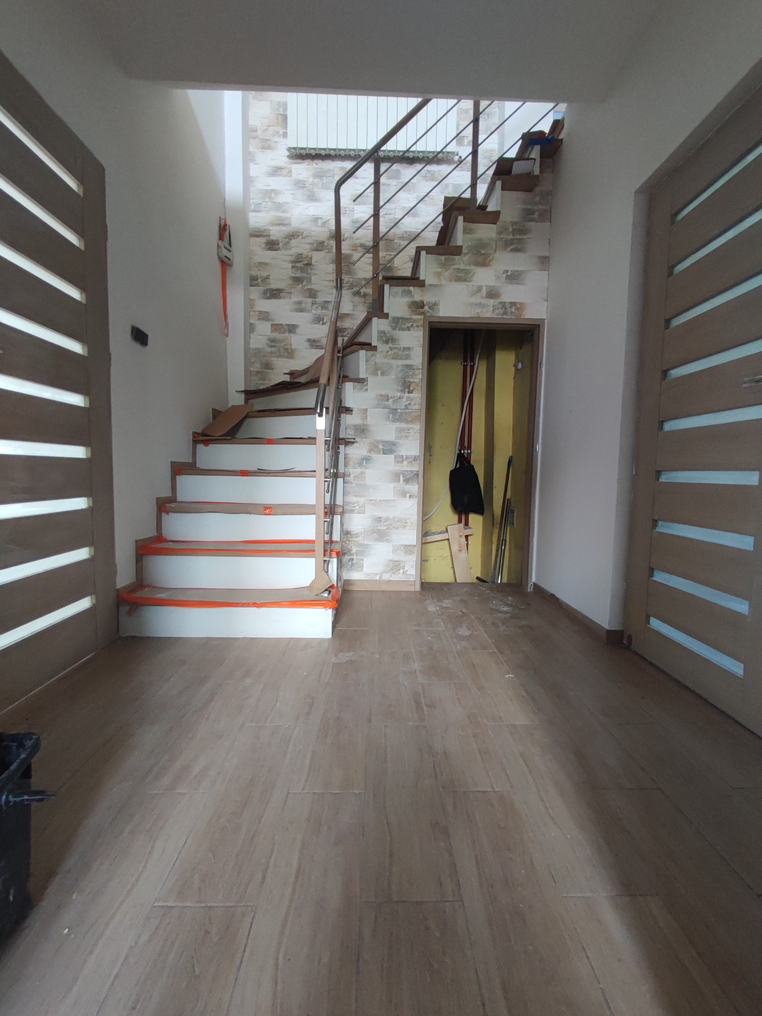 Wnętrze w trakcie prac wykończeniowych: drewnopodobna podłoga, schody z zabezpieczonymi stopniami prowadzące na górę, ściana wyłożona dekoracyjną cegłą, widoczne drzwi z poziomymi przeszkleniami...