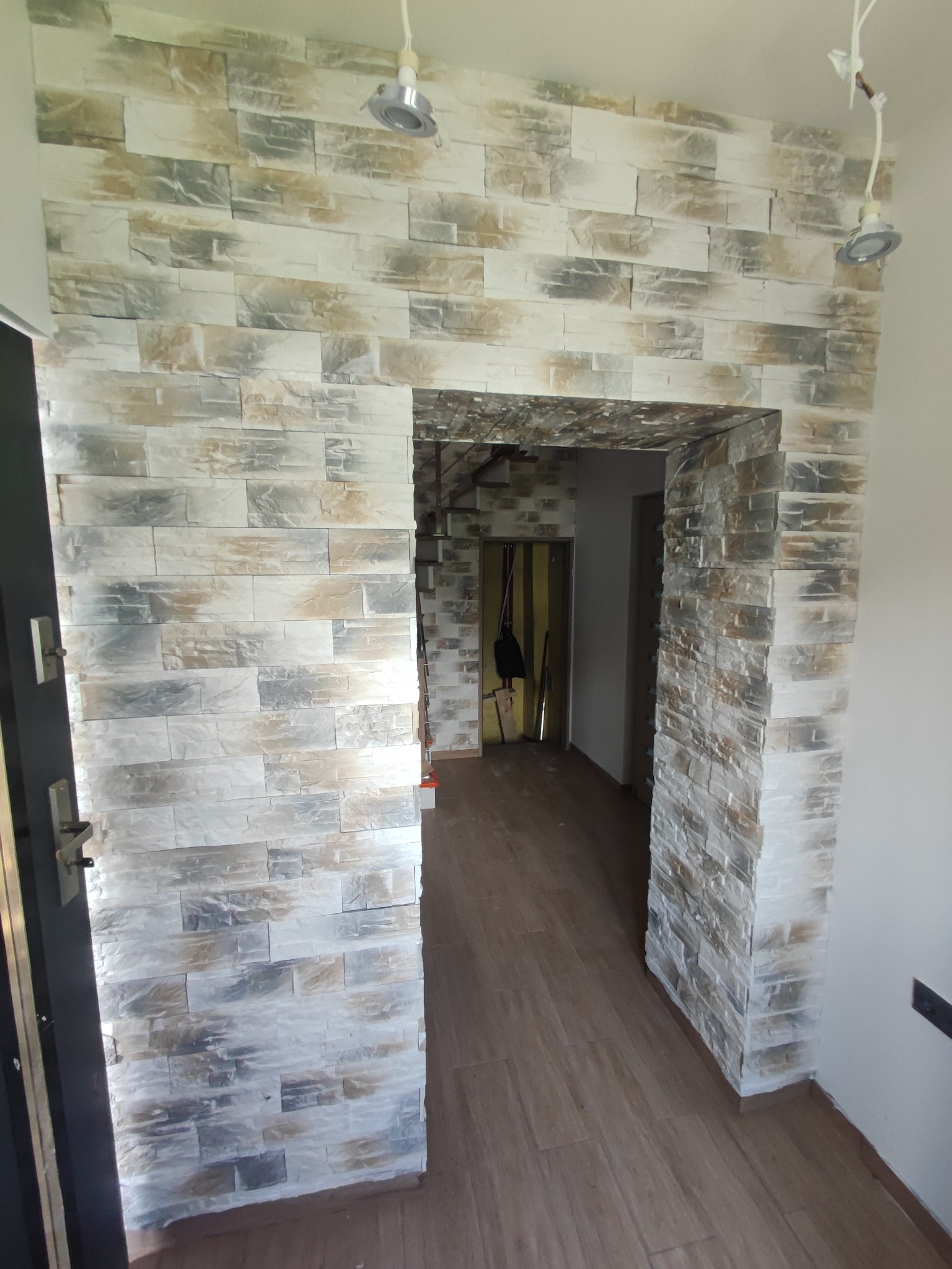 Wykończone przejście w mieszkaniu z dekoracyjną ścianą z paneli imitujących kamień w odcieniach szarości i beżu, z widocznymi punktowymi lampami sufitowymi i drewnopodobną podłogą.