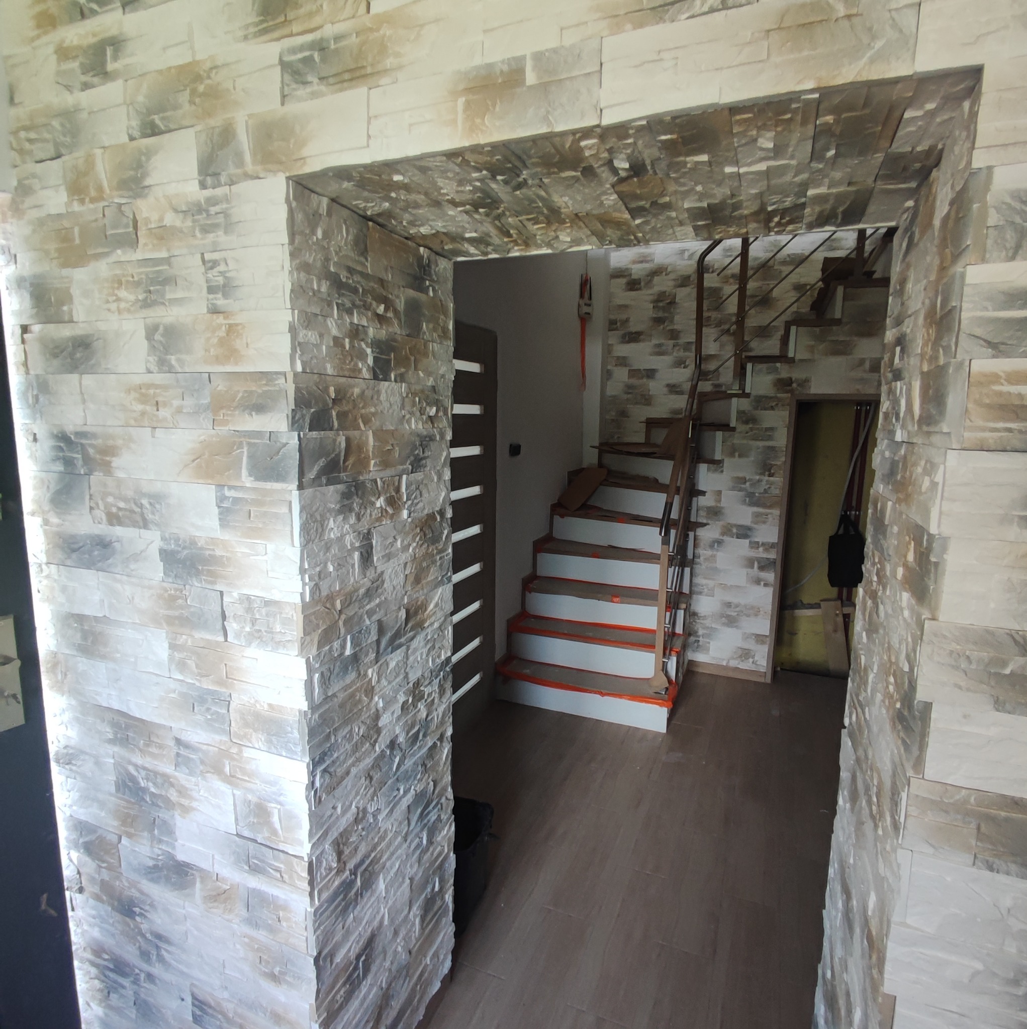 Wykończone przejście z dekoracyjną okładziną kamienną w odcieniach beżu i szarości, widoczne schody z zabezpieczonymi stopniami oraz częściowo wykończona ściana w tle.