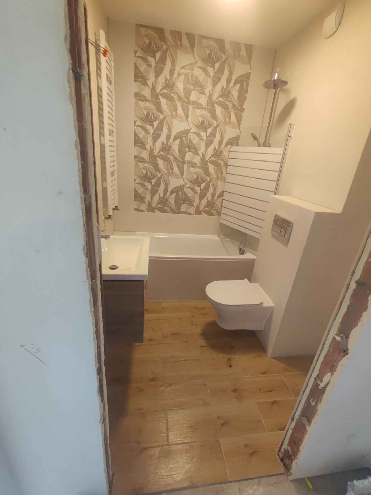 Wnętrze łazienki w trakcie remontu, widoczne elementy instalacji, płytki imitujące drewno, umywalka z szafką, sedes podwieszany i grzejnik drabinkowy na ścianie z tapetą w motywy roślinne.