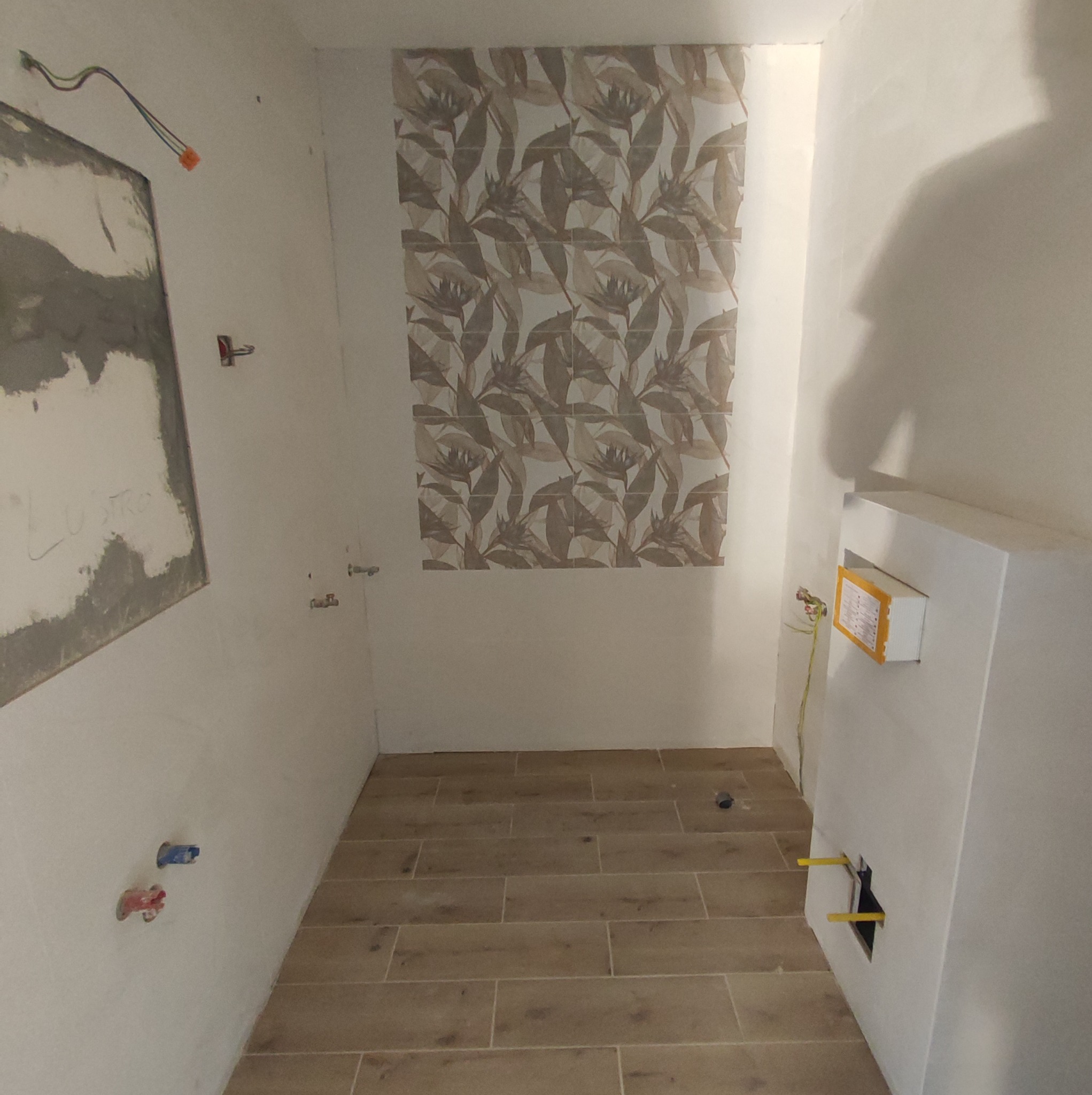 Wnętrze łazienki w trakcie remontu: częściowo położone płytki z motywem liści na ścianie, drewnopodobna podłoga, przygotowane instalacje wodne i elektryczne, ściana z napisem 'LUSTRO' przed...