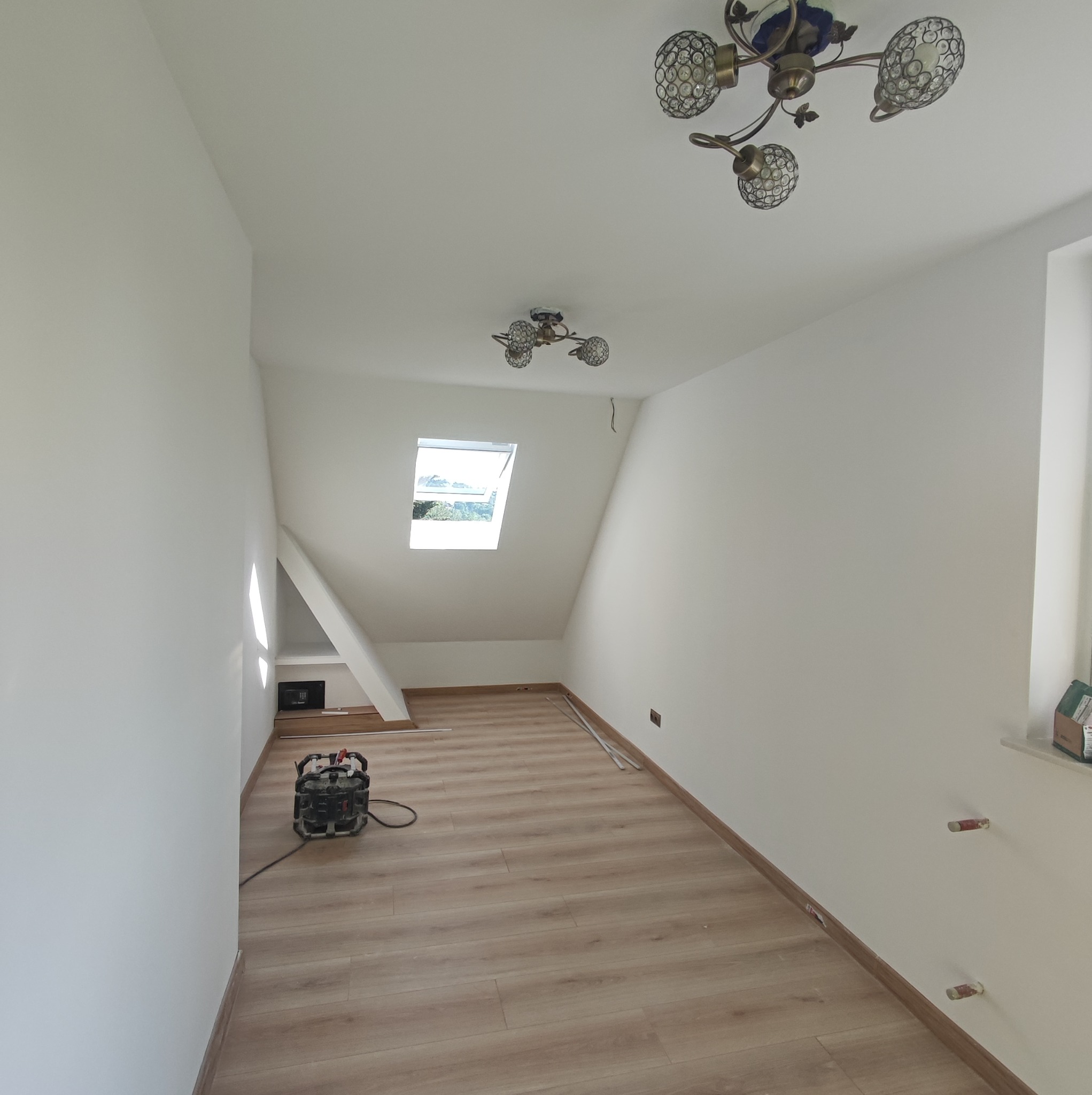 Poddasze po remoncie: świeżo pomalowane białe ściany, ułożona jasna podłoga laminowana, widoczne narzędzia i okno dachowe.