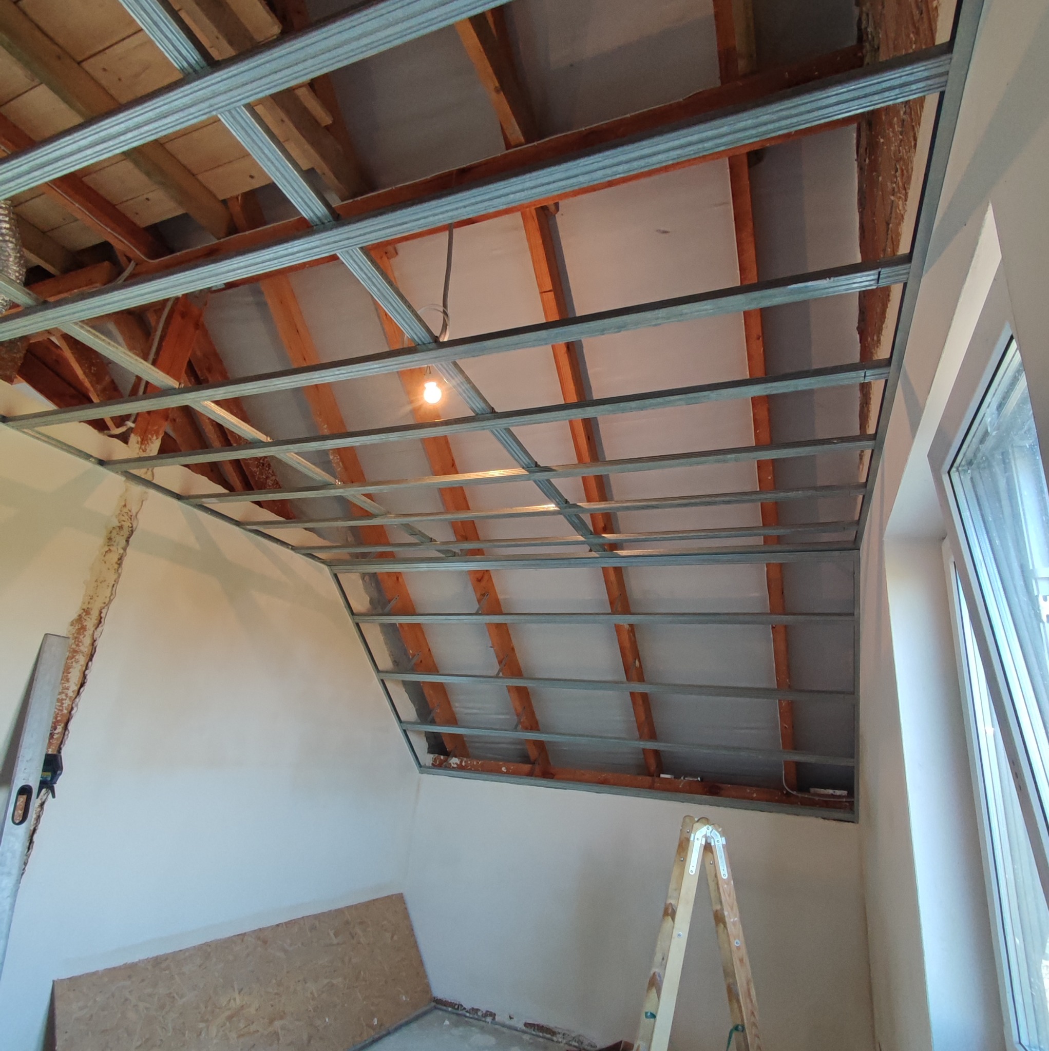 Montaż metalowej konstrukcji podwieszanego sufitu, widoczne drewniane elementy stropu, oświetlenie tymczasowe, drabina oparta o ścianę z oknem.