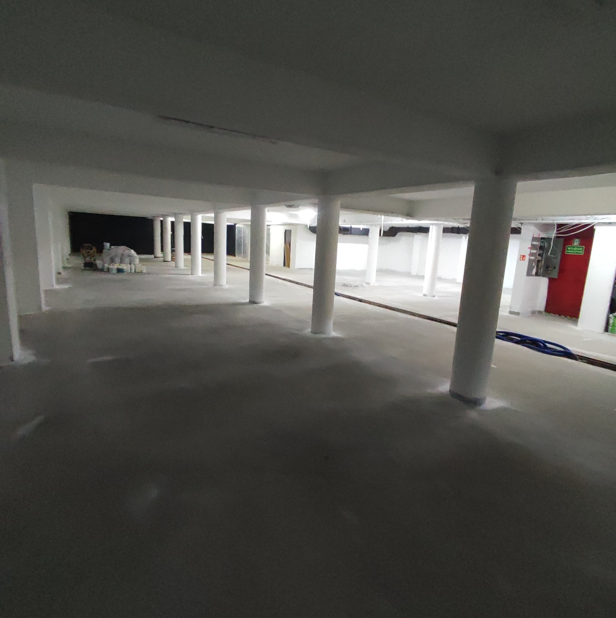 Surowe, puste wnętrze biurowe w trakcie prac wykończeniowych, z kolumnami, świeżo wylaną posadzką i przygotowanymi materiałami budowlanymi w tle.