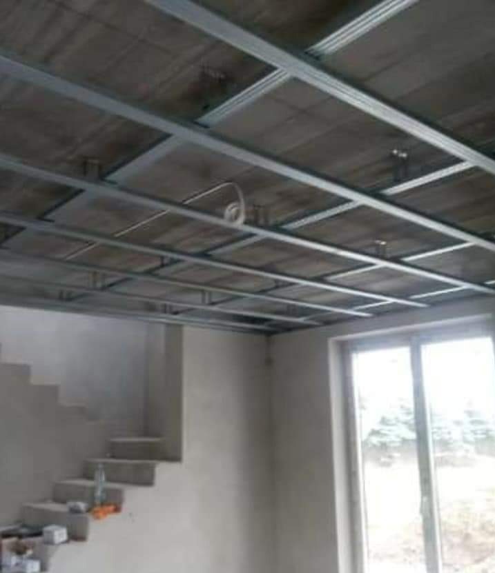 Metalowa konstrukcja podwieszanego sufitu zamontowana w surowym pomieszczeniu z betonowym sufitem, widoczne schody i okno.