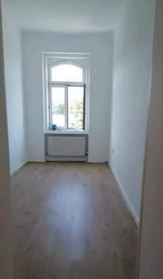 Pusty pokój po remoncie z jasnymi ścianami, drewnianą podłogą i oknem z widokiem na zieleń.