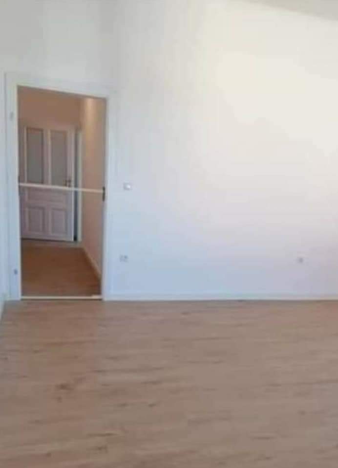 Puste, jasne pomieszczenie po remoncie: świeżo pomalowane białe ściany, jasnobrązowa podłoga imitująca drewno, widok na białe drzwi w głębi.