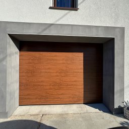 Vindue - Zamknięta brama garażowa segmentowa w kolorze drewna, otoczona szarą, betonową ramą, na tle białej fasady domu z małym oknem na górze.