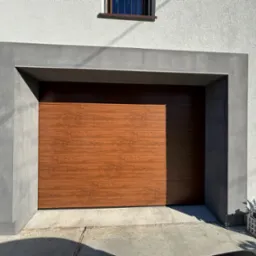 Zamknięta brama garażowa segmentowa w kolorze drewna, otoczona szarą, betonową ramą, na tle białej fasady domu z małym oknem na górze.