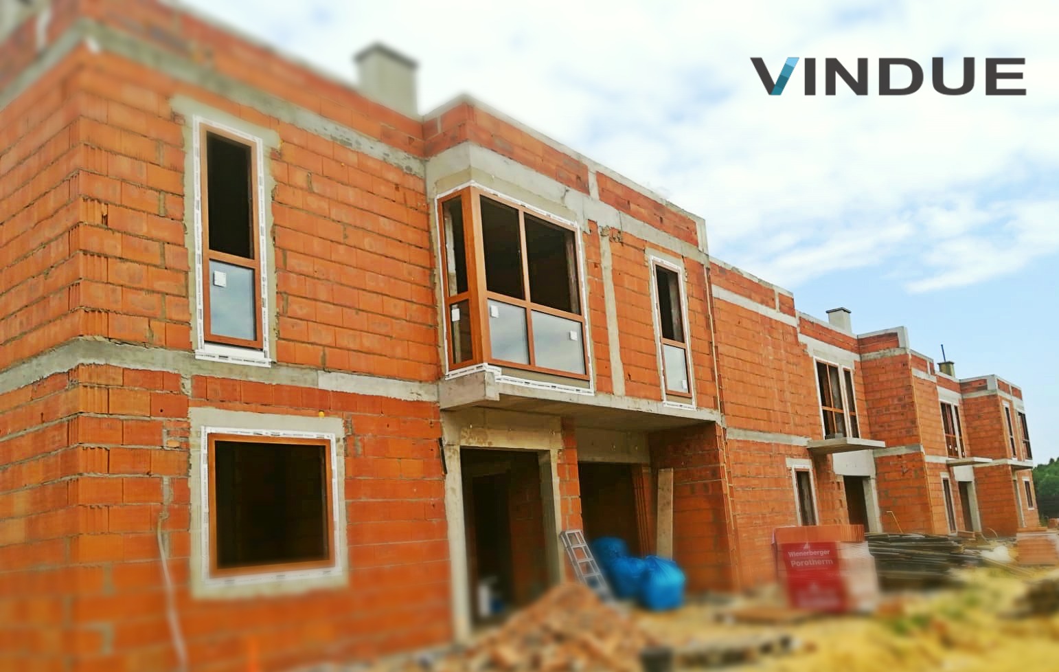Budynek w trakcie budowy z zamontowanymi oknami z brązowymi ramami, widoczne materiały budowlane i logo firmy VINDUE na górze zdjęcia.