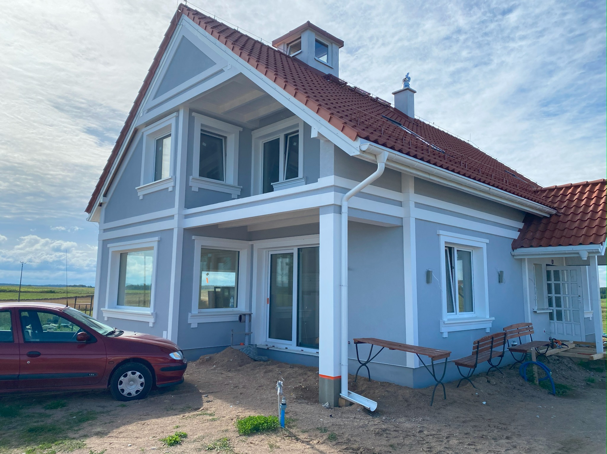 Niebieski dom jednorodzinny z białymi ramami okiennymi i drzwiami przesuwnymi, widoczny czerwony samochód i tymczasowe ławki na zewnątrz, teren w trakcie prac wykończeniowych.