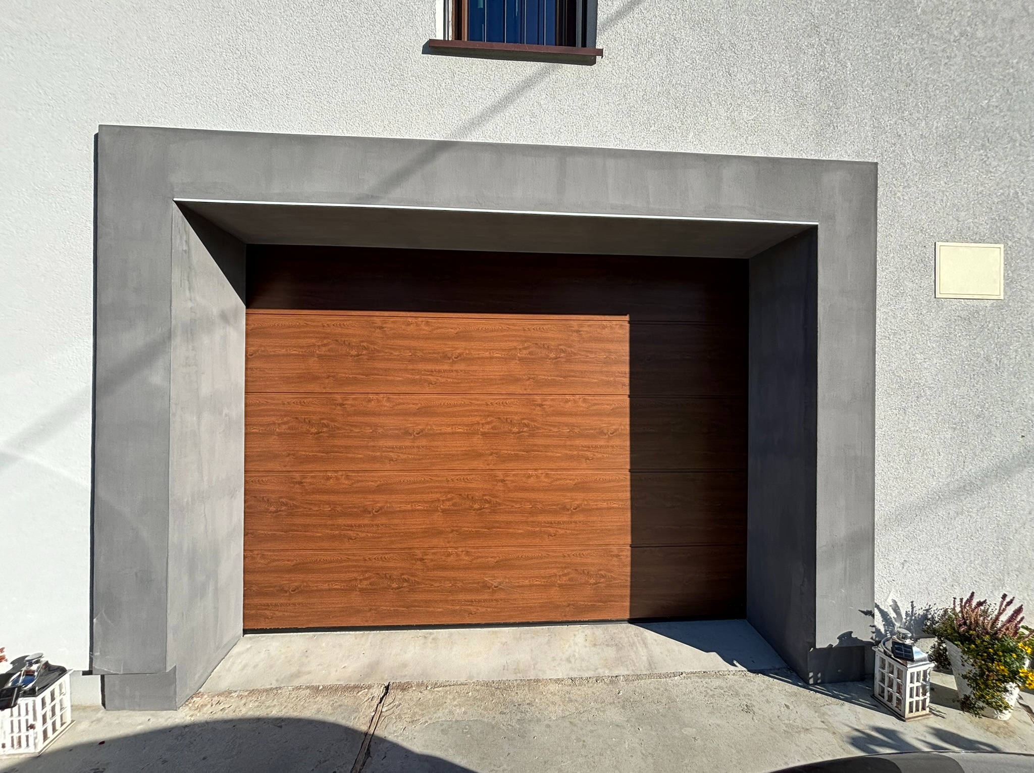 Zamknięta brama garażowa segmentowa w kolorze drewna, otoczona szarą, betonową ramą, na tle białej fasady domu z małym oknem na górze.