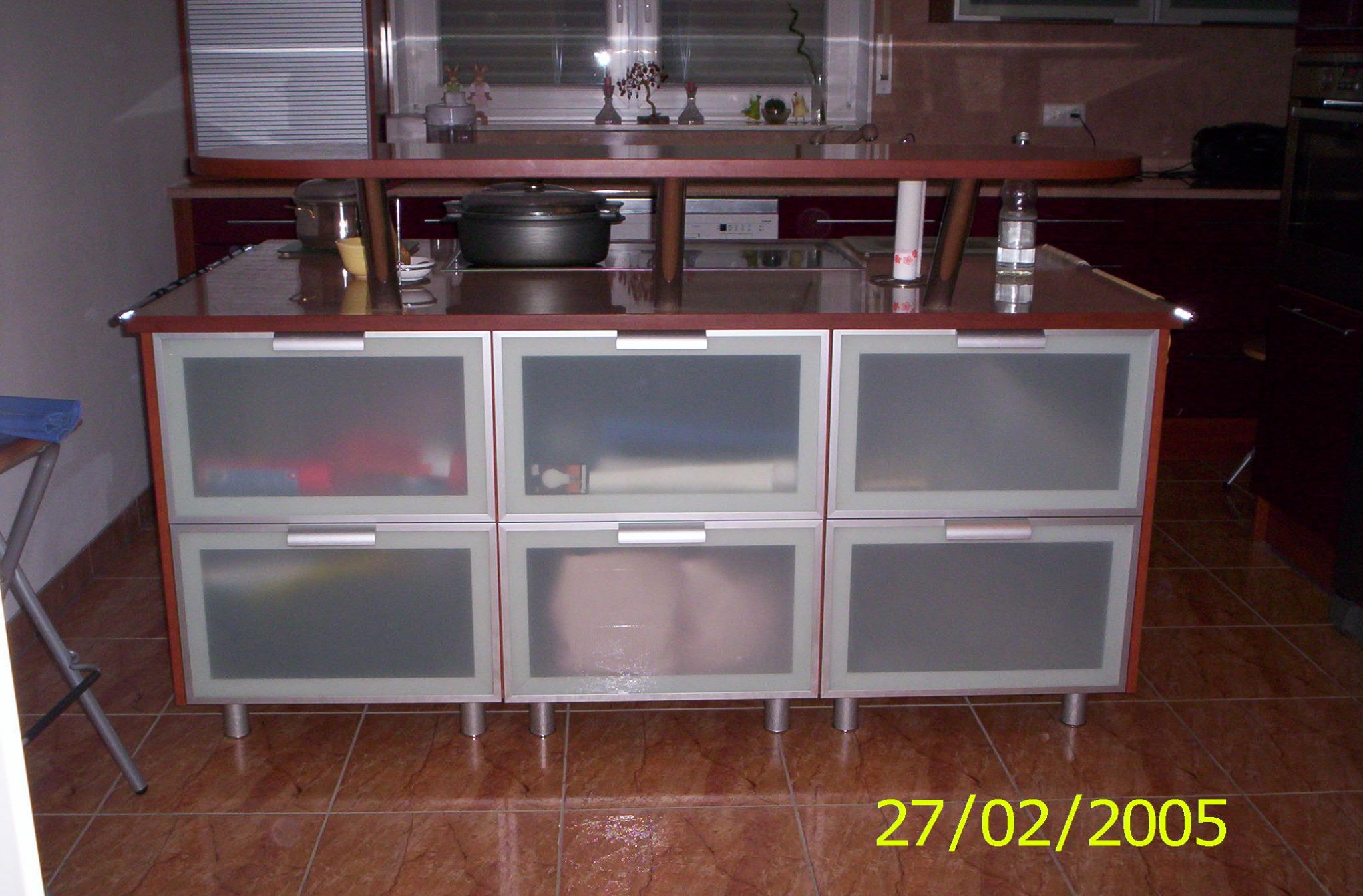 Kuchenna szafka z sześcioma szufladami z matowego szkła, na blacie garnki i akcesoria kuchenne, widok na okno z roletami i dekoracjami.