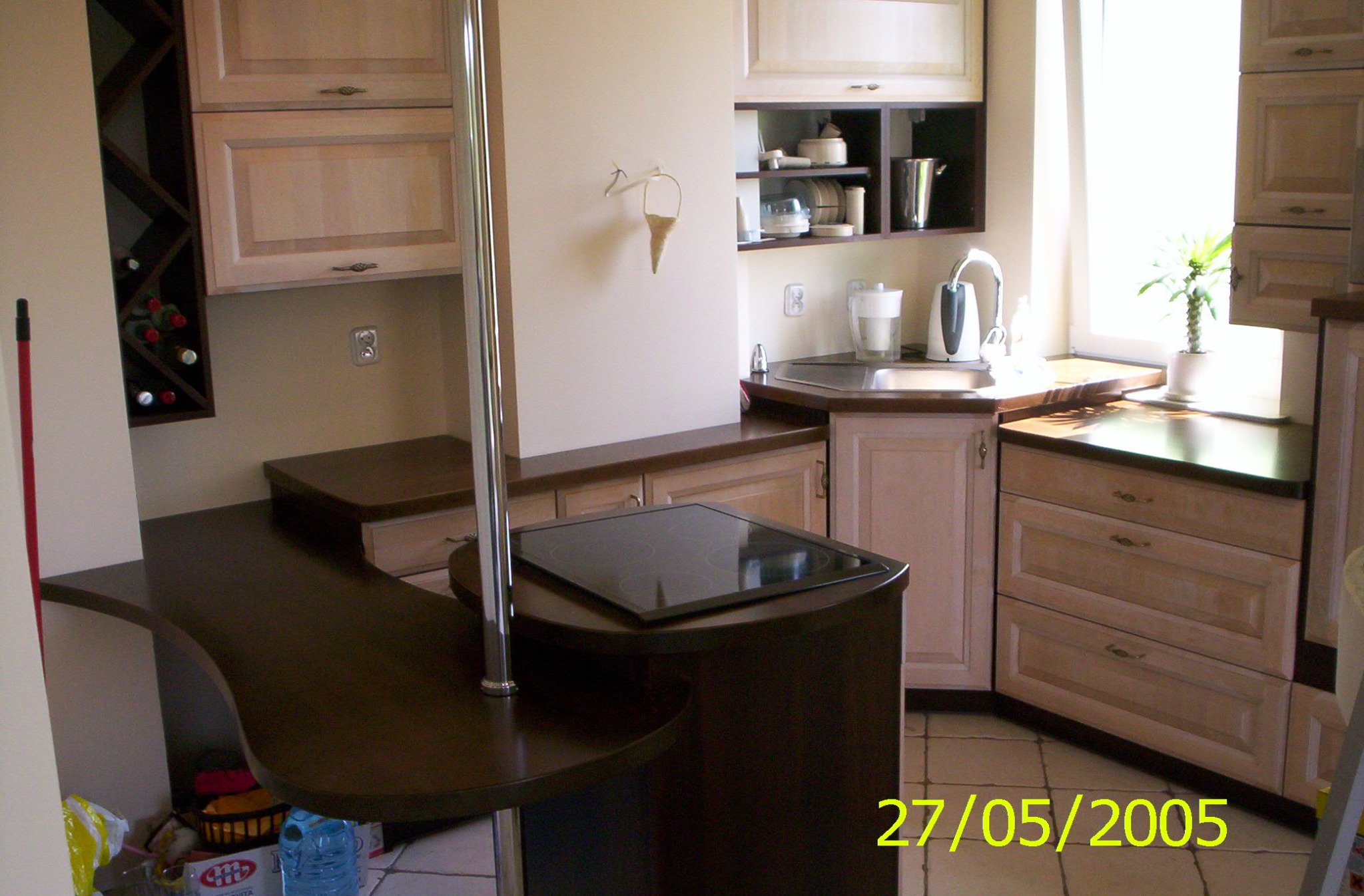 Jasna kuchnia z barkiem w Szczecinie, widok na szafki z drewnianymi frontami, blat z ciemnego drewna i płytę indukcyjną. Na ścianie wisi ozdobny wazonik. W tle widać okno z rośliną.