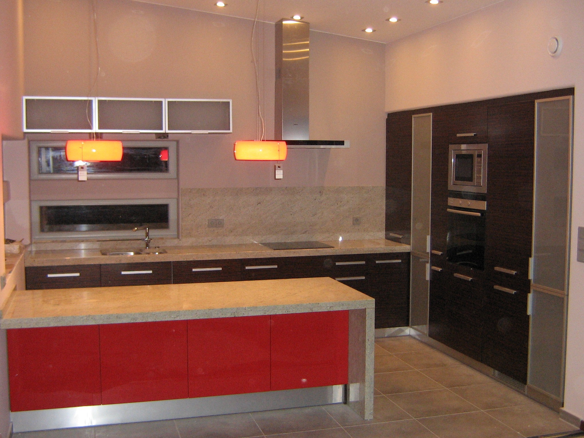Nowoczesna kuchnia z wyspą w kolorze czerwieni, ciemne drewniane szafki, granitowy blat, stalowy okap i podwieszane lampy z pomarańczowym światłem.