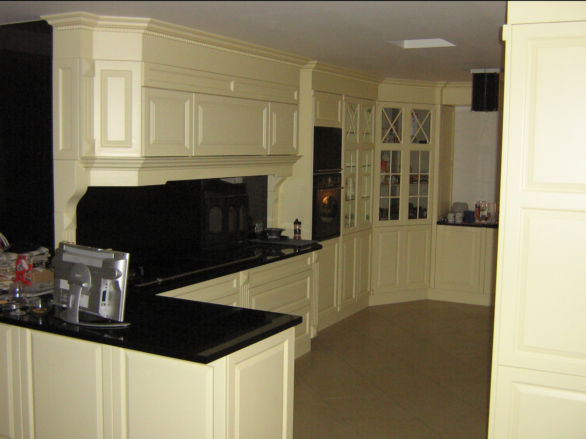 Klasyczna kuchnia w odcieniach kremu z czarnym blatem, widoczny monitor na blacie i przeszklone szafki z dekoracyjnymi krzyżami.