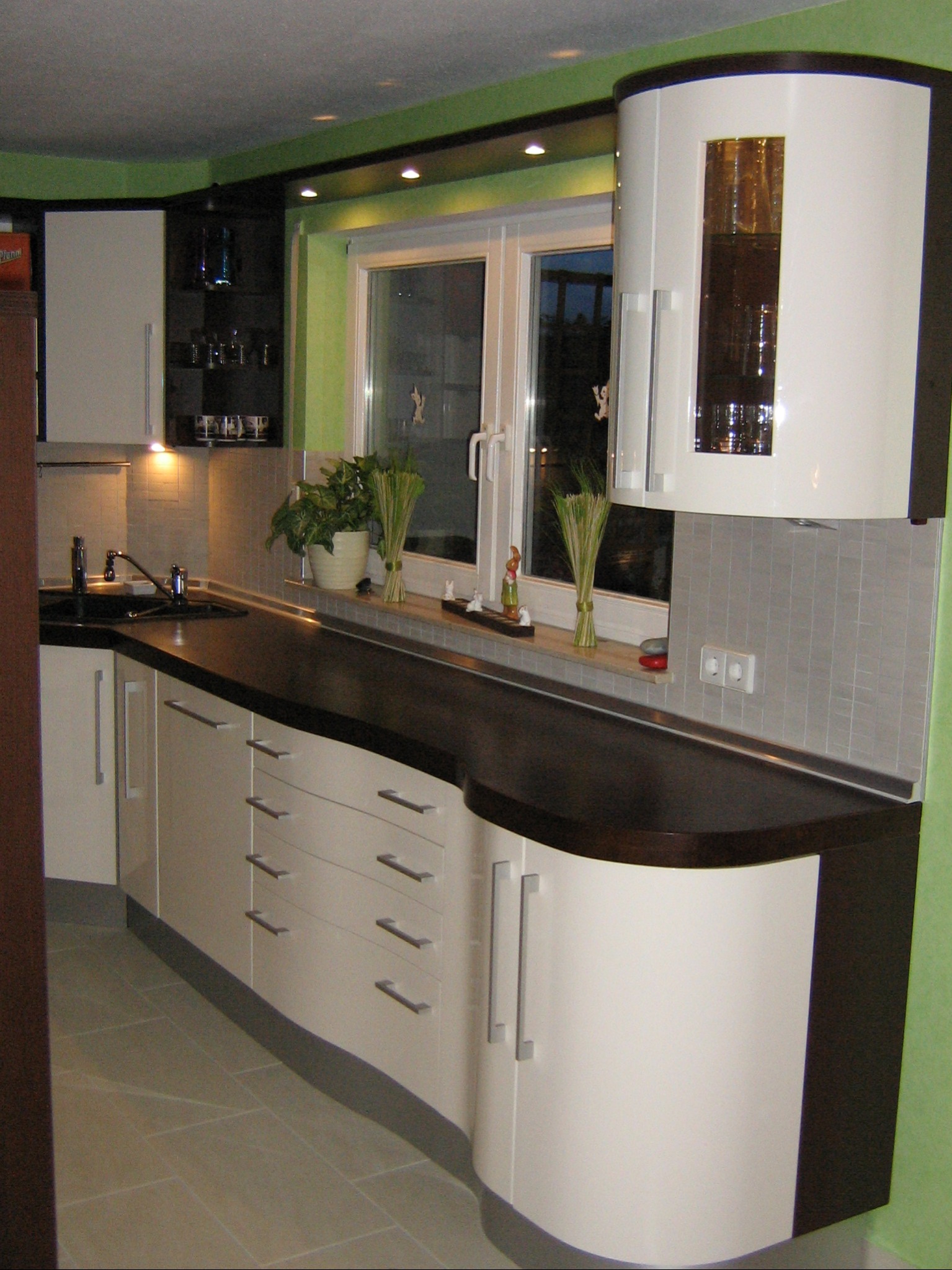 Nowoczesna kuchnia z białymi szafkami z zaokrąglonymi narożnikami, ciemnym blatem i szarymi płytkami nad zlewem, z oknem z roślinami i dekoracjami.